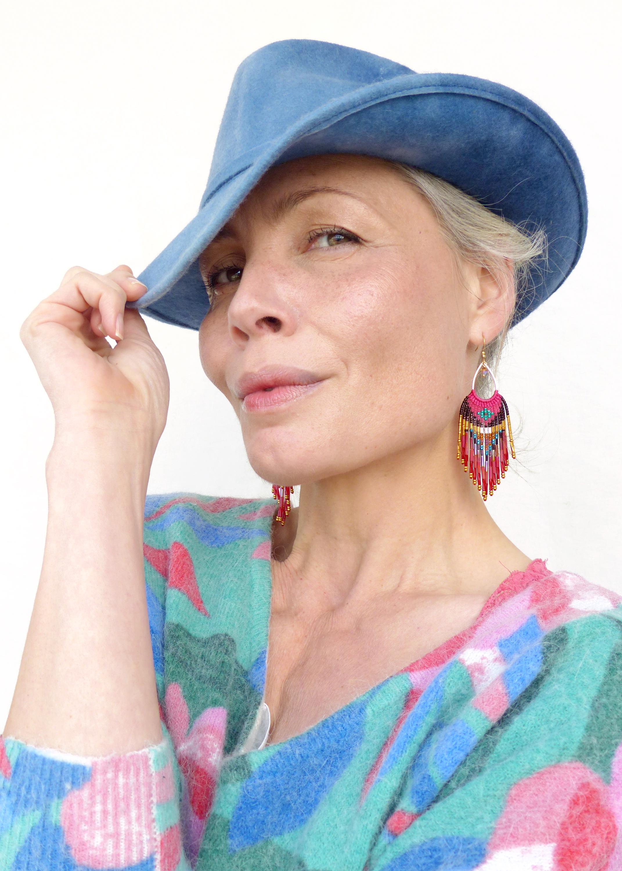 Peut inclure: Une personne portant un chapeau bleu clair et des boucles d'oreilles à perles colorées. Les boucles d'oreilles ont une forme de goutte d'eau avec des franges de perles roses, rouges et dorées. Elle porte un pull avec un motif floral dans les tons de bleu, vert et rose.