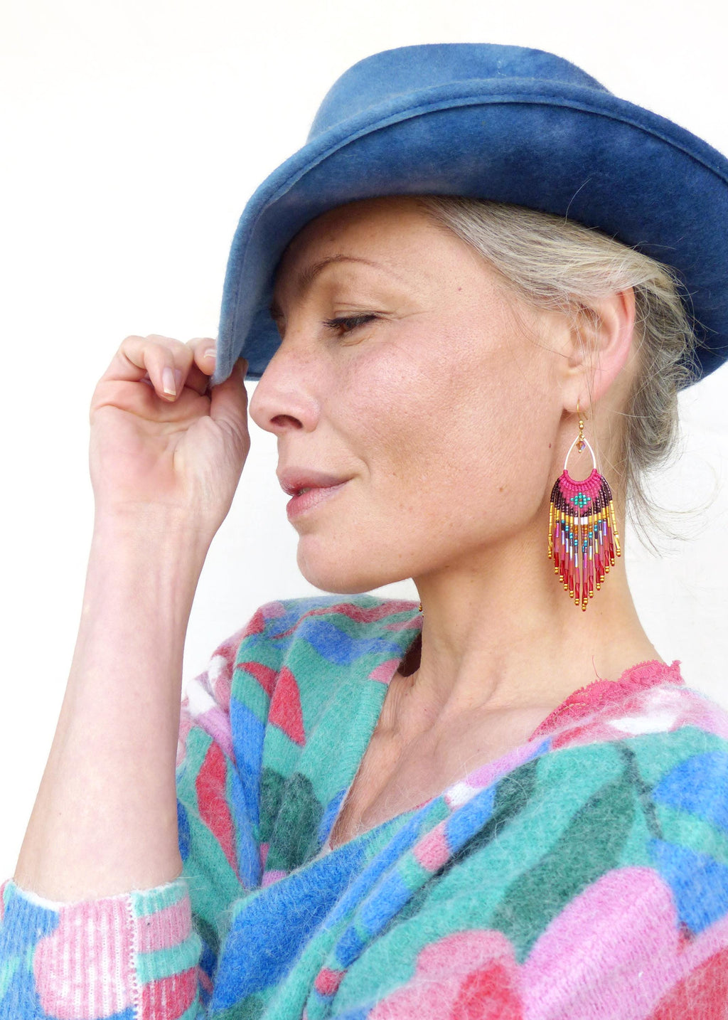 Peut inclure: Une femme porte un chapeau bleu et des boucles d'oreilles en perles colorées. Les boucles d'oreilles présentent un motif rose, rouge, jaune et bleu avec des accents dorés. Elle porte également un pull à motifs roses, bleus et verts.