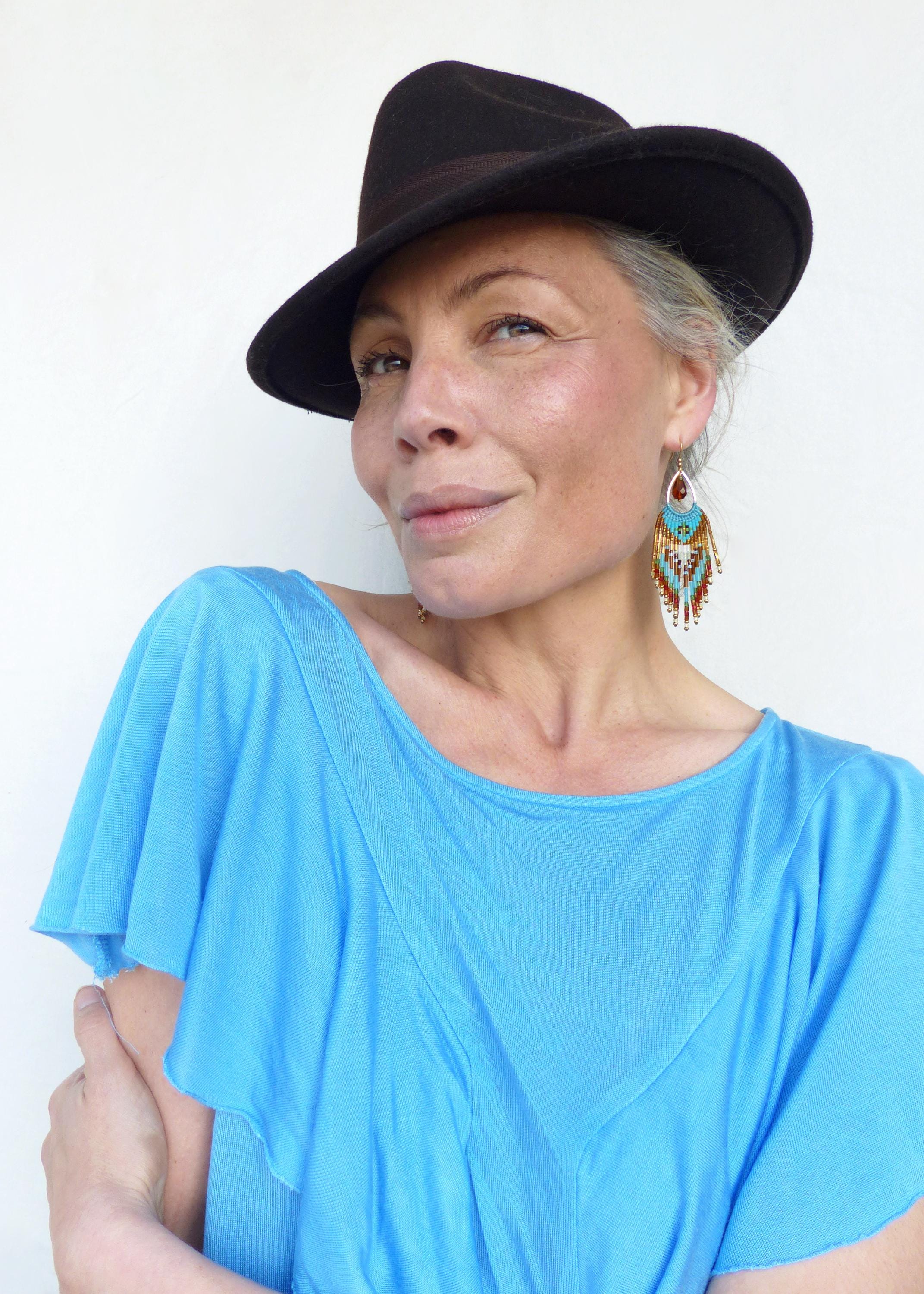 Peut inclure: Une femme porte un chapeau fedora marron foncé et un haut bleu turquoise. Elle porte également des boucles d'oreilles en perles avec des accents turquoise, marron et blancs. Le fond est un mur blanc uni.