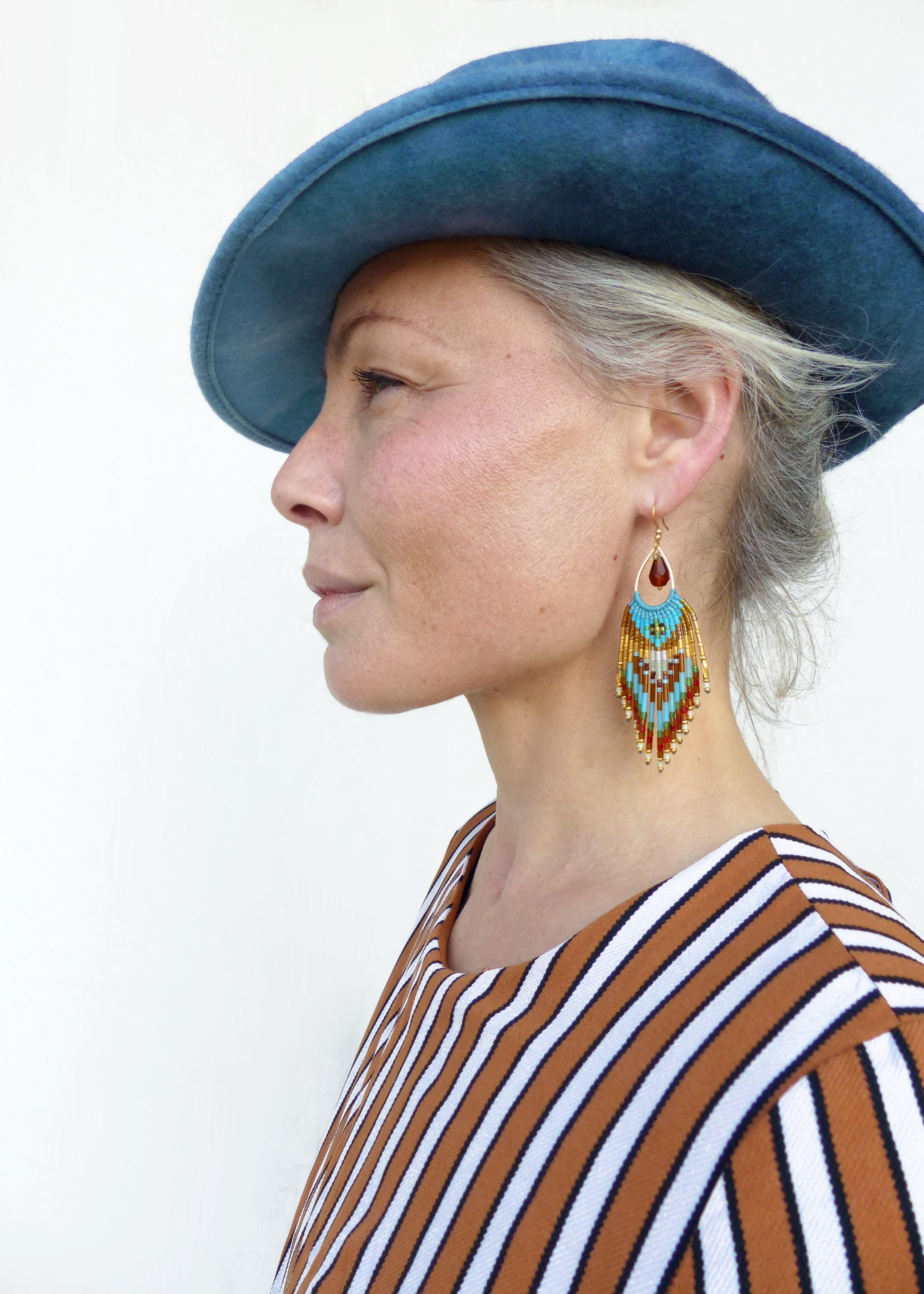 Peut inclure: Une femme porte un chapeau bleu et un haut rayé marron et blanc. Elle porte de grandes boucles d'oreilles en perles colorées avec une pierre rouge en forme de goutte d'eau. Les boucles d'oreilles sont turquoise, dorées et marron.
