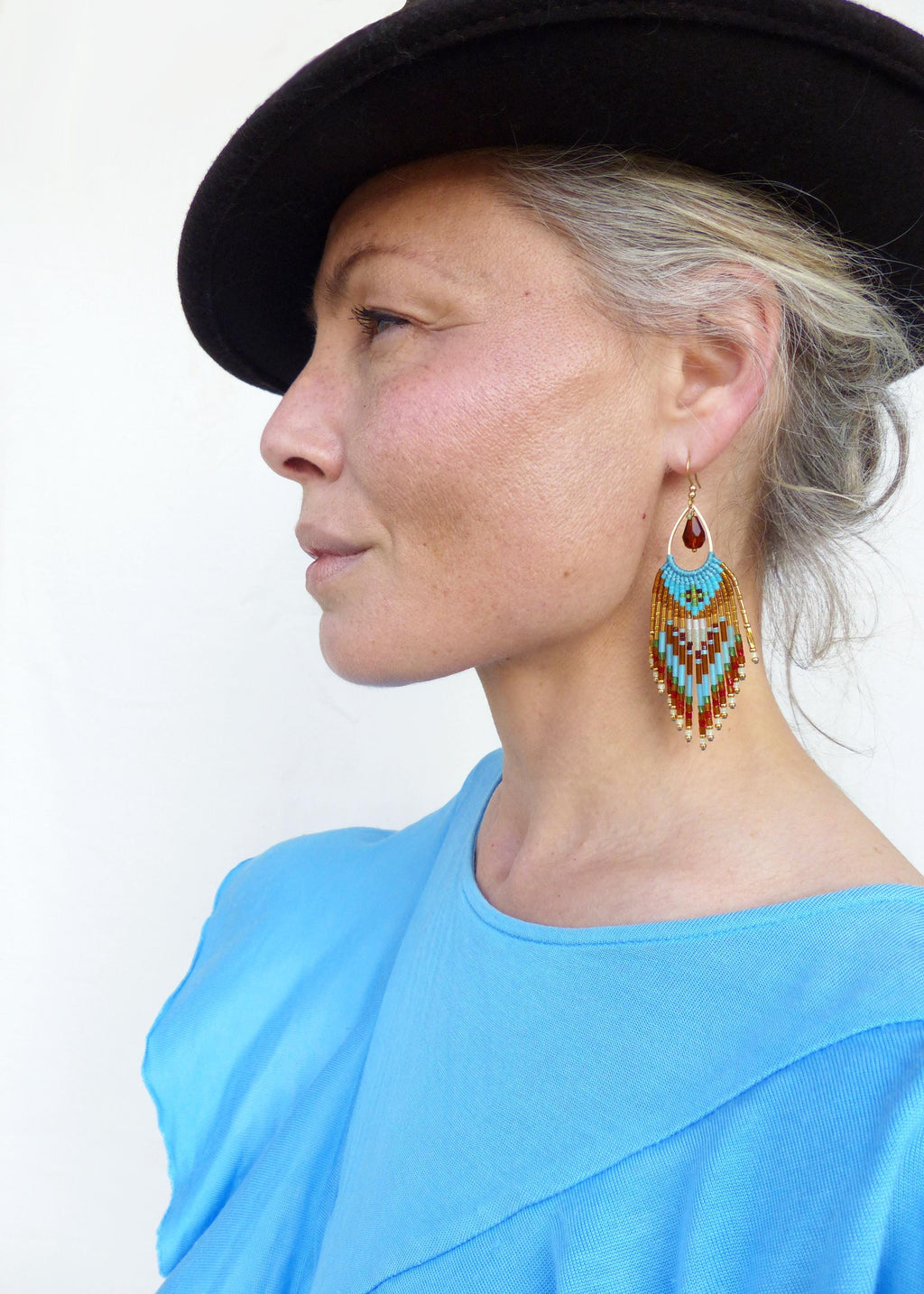Peut inclure: Une femme porte un chapeau noir et un haut bleu clair. Elle porte de longues boucles d'oreilles perlées avec une pierre rouge en forme de goutte d'eau en haut. Les boucles d'oreilles ont un motif turquoise, doré et rouge.