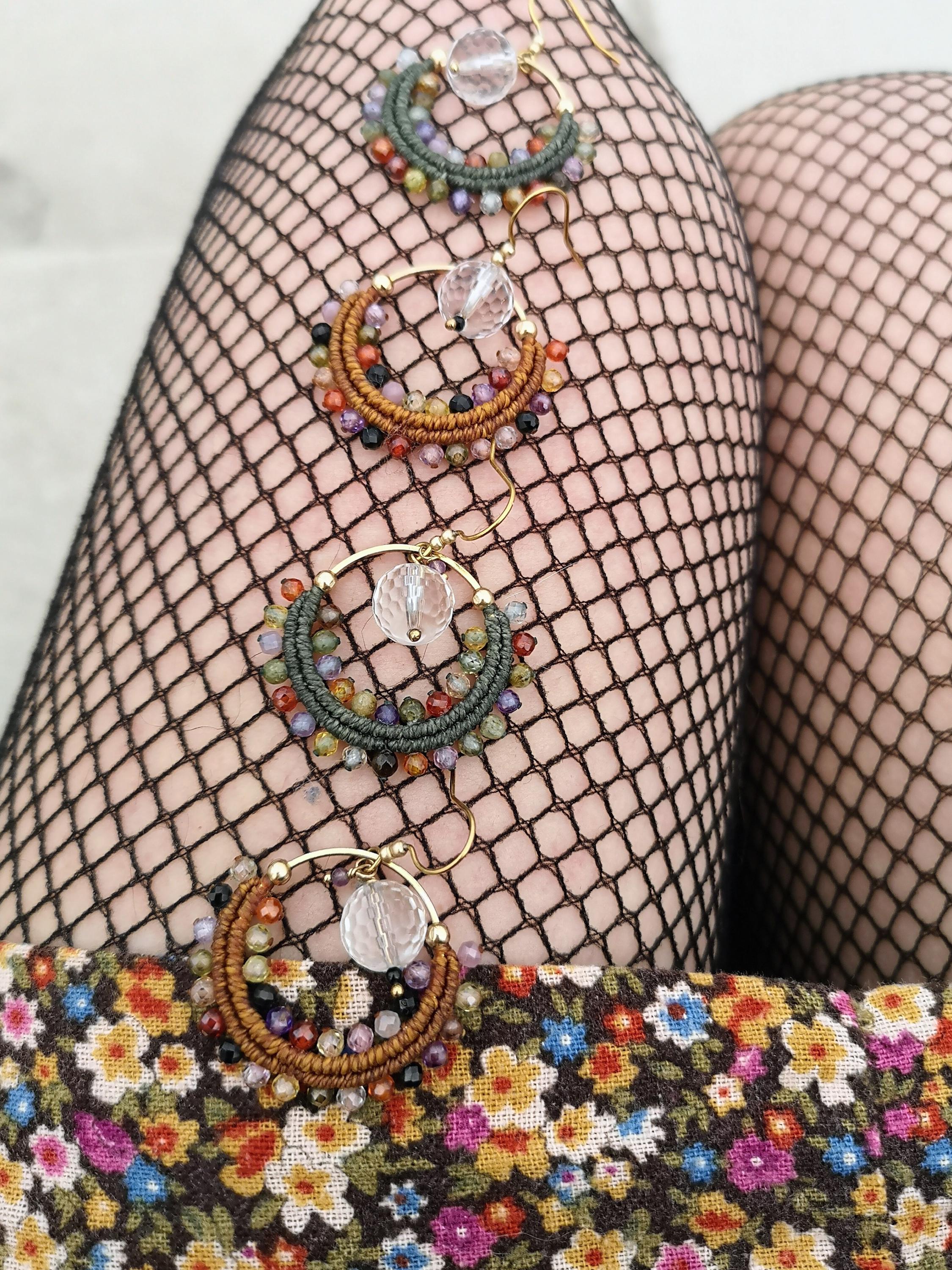Peut inclure: Quatre paires de boucles d'oreilles créoles avec un design macramé. Les créoles sont ornées de perles colorées et d'un cristal transparent. Les boucles d'oreilles sont dorées et ont un aspect artisanal unique. Le fond comprend des bas résille noirs et un tissu floral.