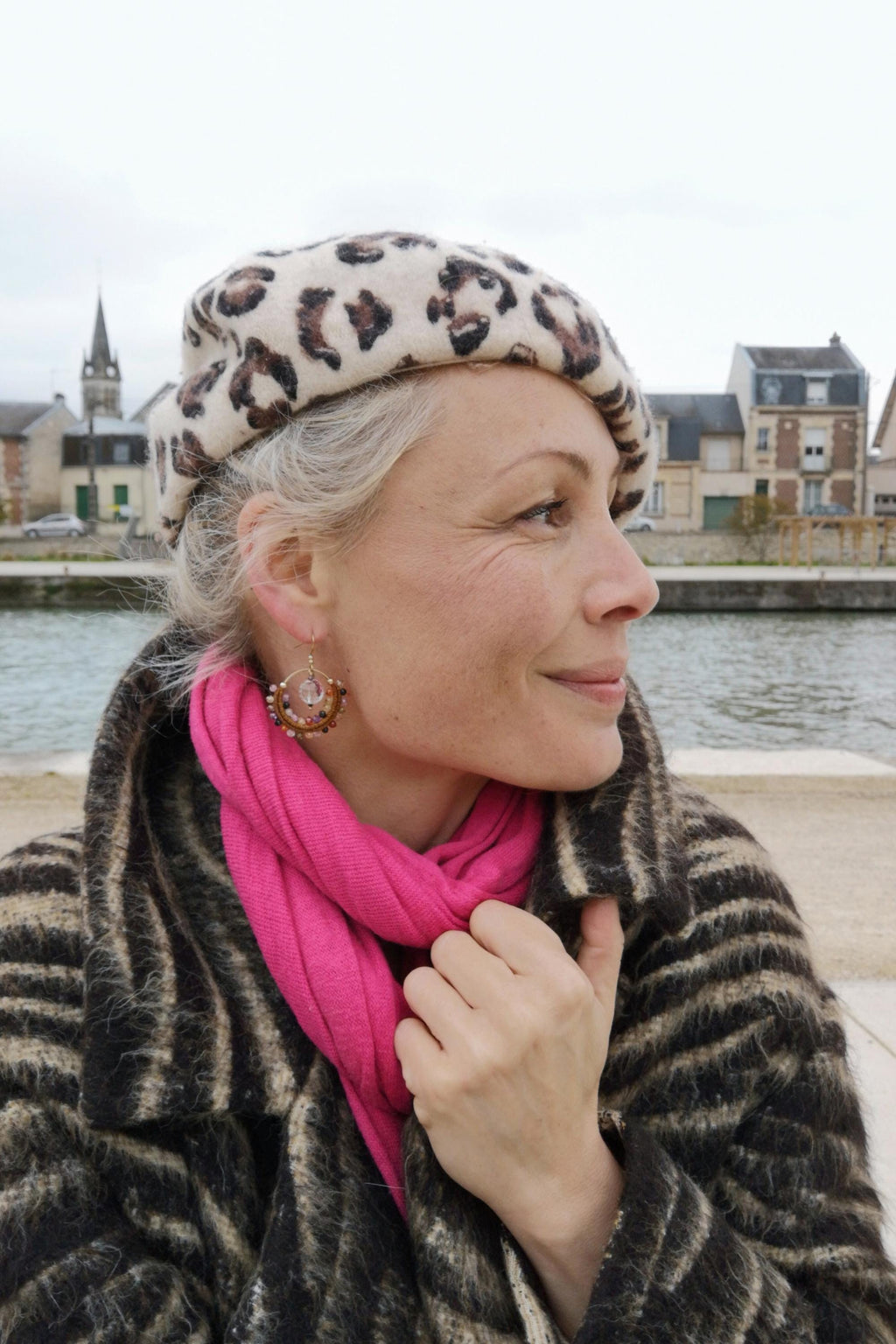 Peut inclure: Une femme porte un béret crème à imprimé léopard marron, une écharpe rose vif et un manteau rayé marron et noir. Les boucles d'oreilles sont dorées avec un motif circulaire et de petites perles. En arrière-plan, un fleuve et des bâtiments.