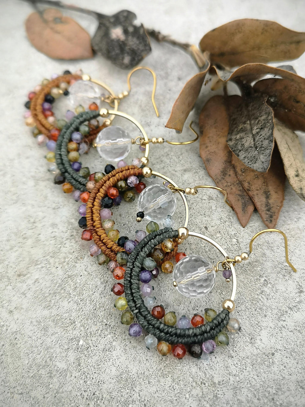 Peut inclure: Boucles d'oreilles créoles dorées avec des accents de perles colorées. Chaque boucle d'oreille présente un cerceau doré avec une perle de cristal transparente et un motif en forme de croissant enveloppé de fil marron et vert, orné de petites perles multicolores.