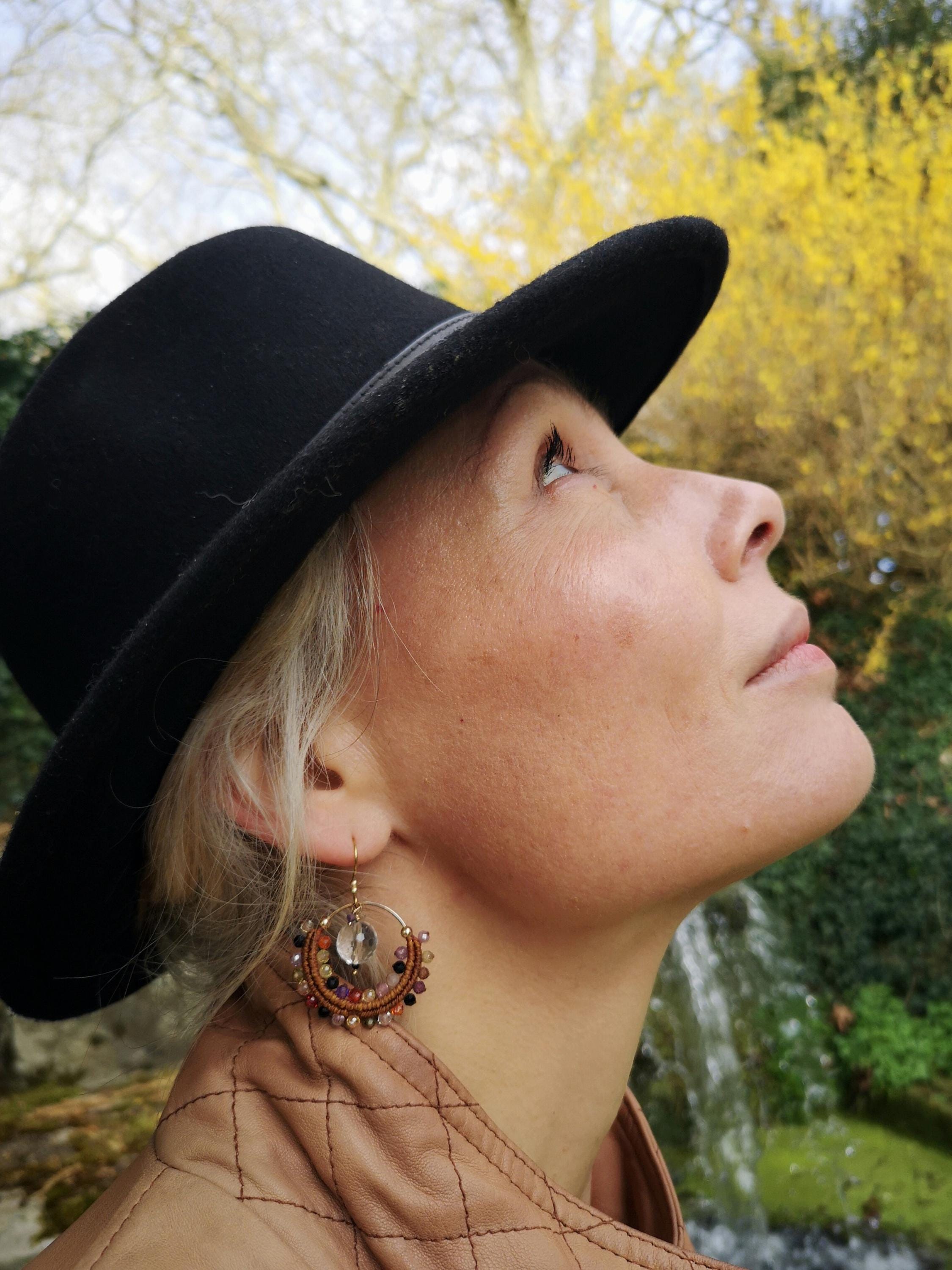 Peut inclure: Une femme porte un chapeau fedora noir et une veste en cuir beige. Elle porte des boucles d'oreilles dorées avec des perles colorées et un cristal transparent. L'arrière-plan présente une cascade et des fleurs jaunes.
