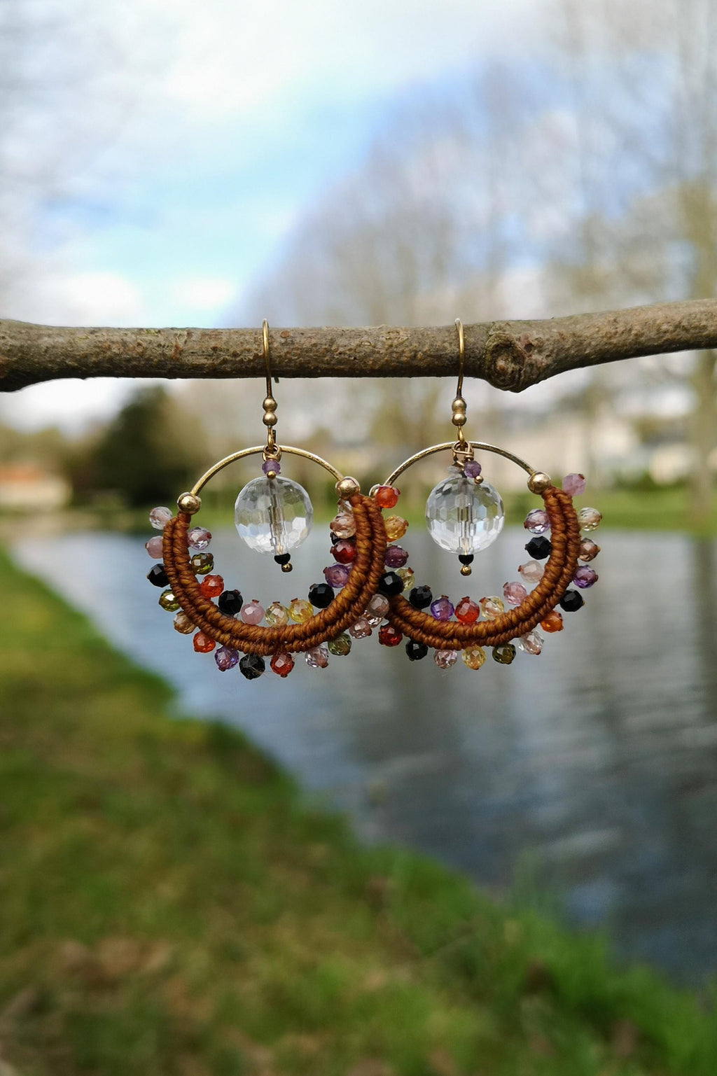 Peut inclure: Une paire de boucles d'oreilles créoles faites à la main, de style bohème. Les boucles d'oreilles sont composées d'un cordon marron tressé, orné de perles colorées dans les tons de rouge, orange, jaune, violet et noir. Chaque boucle d'oreille a une perle de cristal transparente et des accents métalliques dorés.
