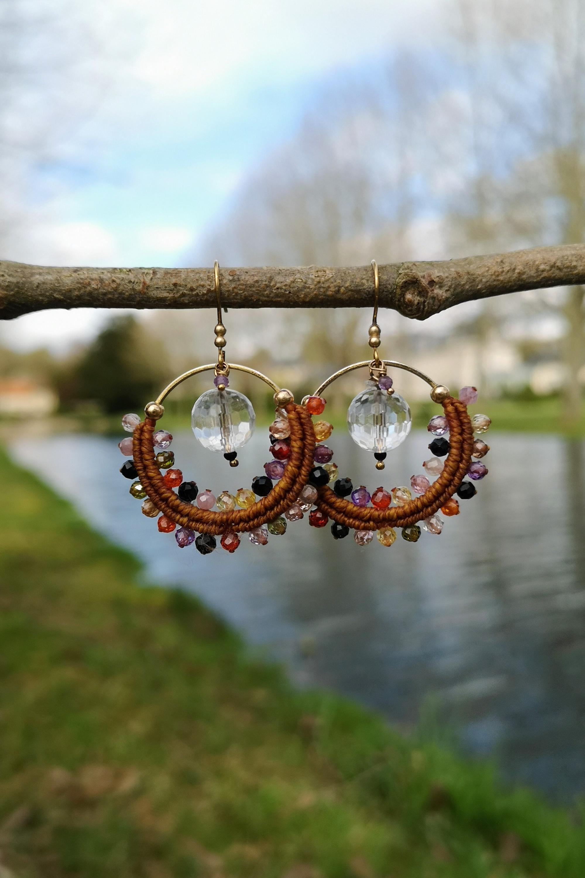 Peut inclure: Une paire de boucles d'oreilles créoles faites à la main, de style bohème. Les boucles d'oreilles sont composées d'un cordon marron tressé, orné de perles colorées dans les tons de rouge, orange, jaune, violet et noir. Chaque boucle d'oreille a une perle de cristal transparente et des accents métalliques dorés.