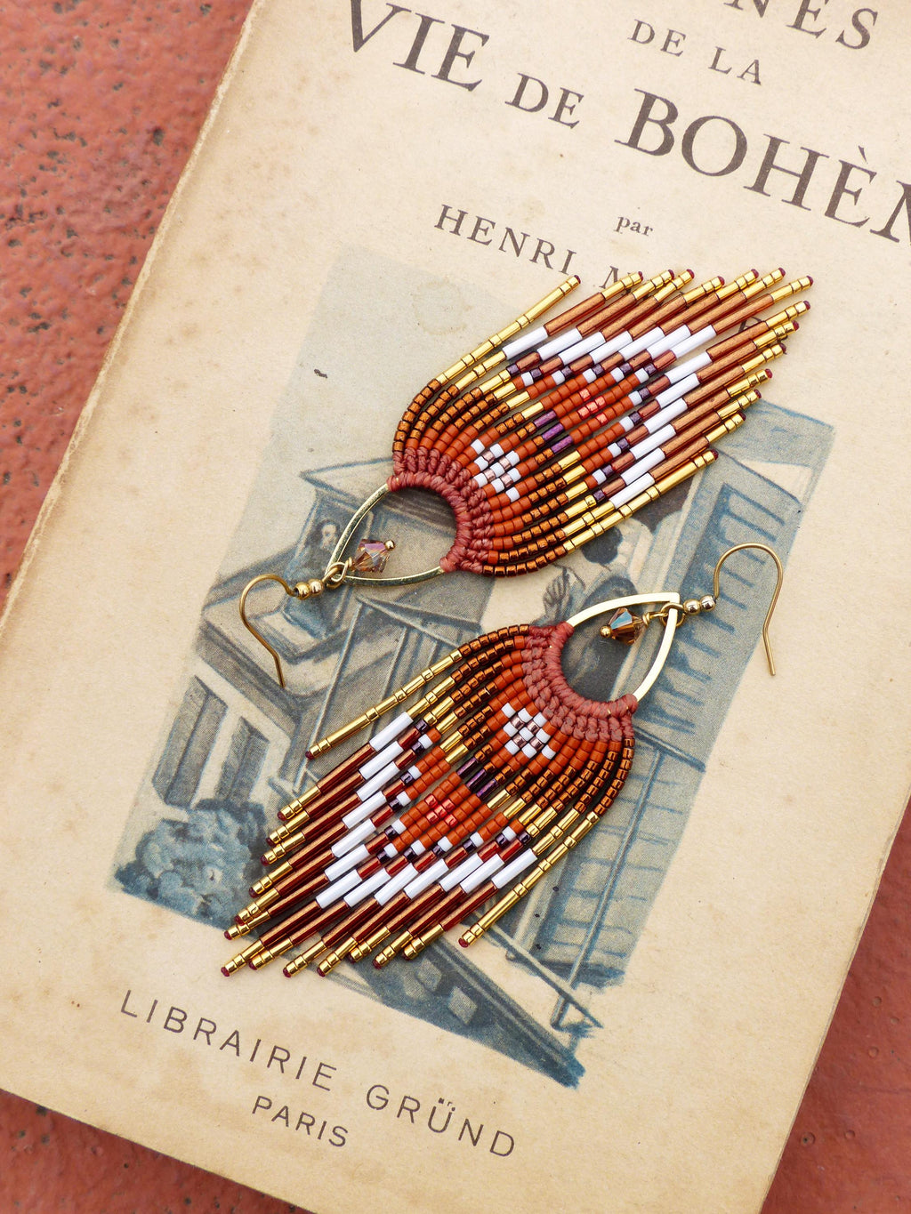 Peut inclure: Une paire de boucles d'oreilles faites à la main avec un design bohème. Les boucles d'oreilles présentent une forme en éventail avec une combinaison de perles rouges, blanches, dorées et marron. Elles sont posées sur une page de livre vintage avec le texte "VIE DE BOHÈME".