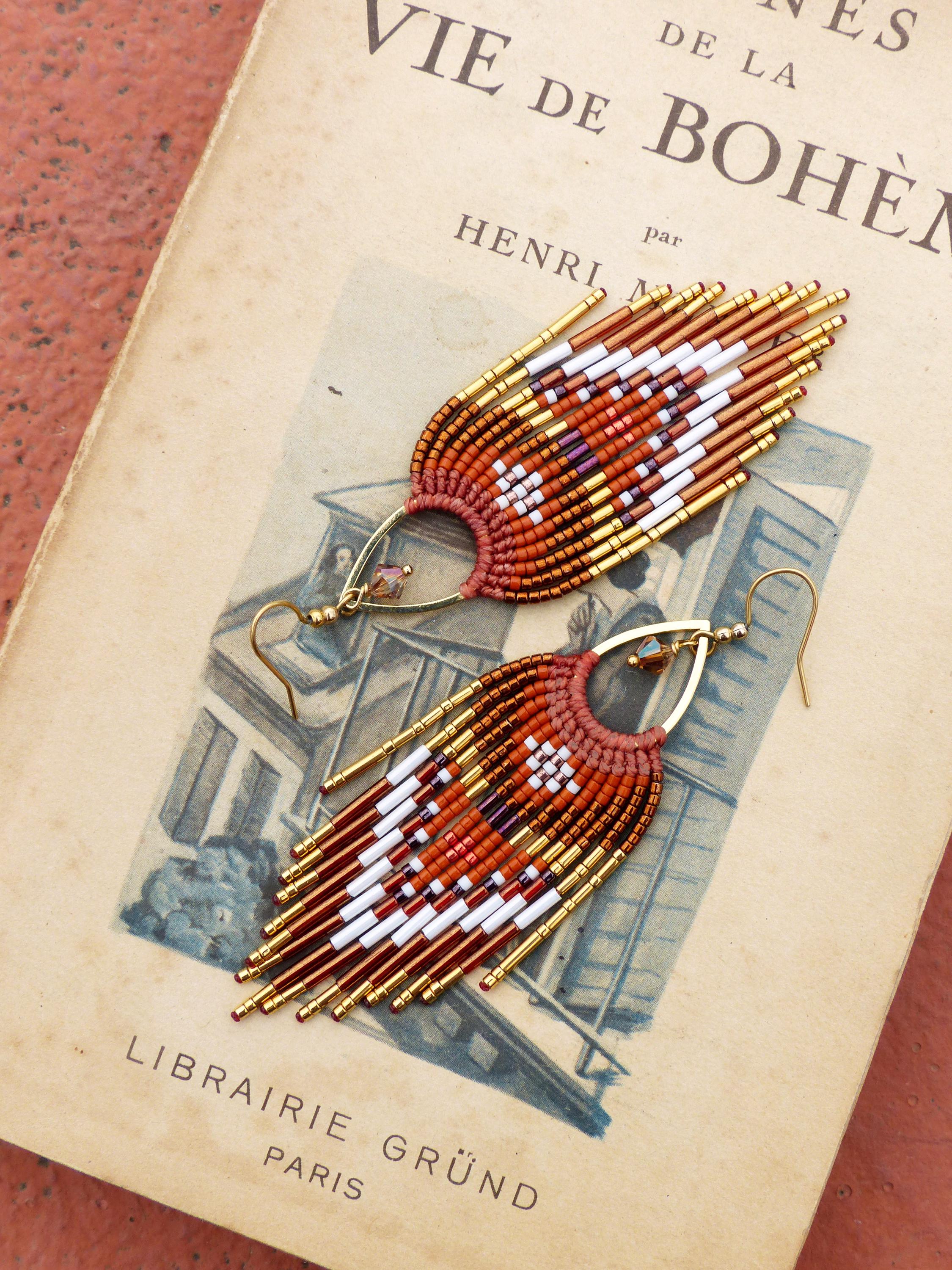 Peut inclure: Une paire de boucles d'oreilles faites à la main avec un design bohème. Les boucles d'oreilles présentent une forme en éventail avec une combinaison de perles rouges, blanches, dorées et marron. Elles sont posées sur une page de livre vintage avec le texte "VIE DE BOHÈME".
