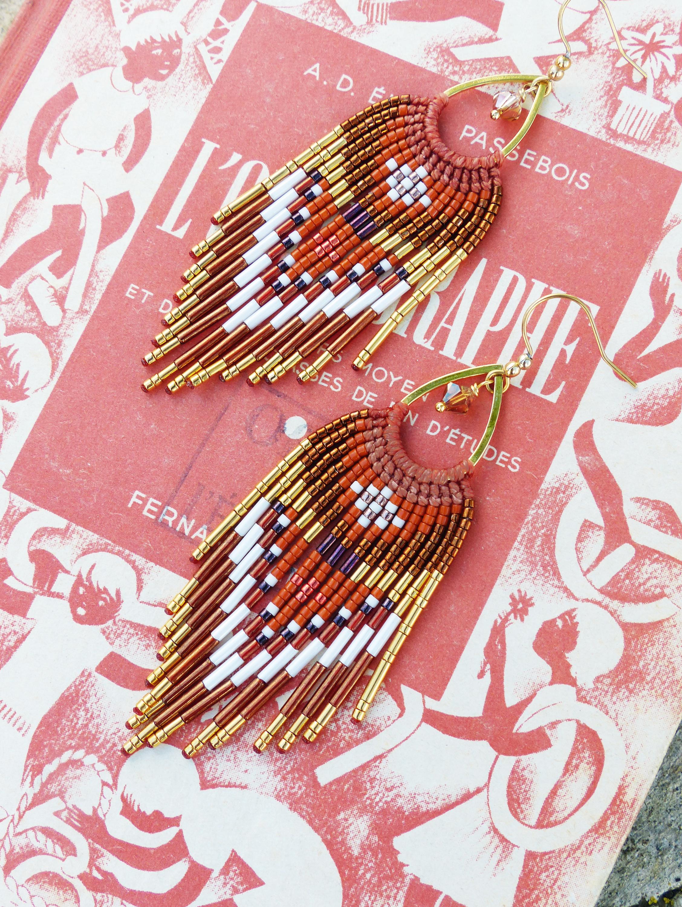Peut inclure: Paire de boucles d'oreilles en perles avec un motif géométrique. Elles présentent des perles orange, blanches, marron et dorées, avec une frange en bas. Le fond est rouge avec du texte.