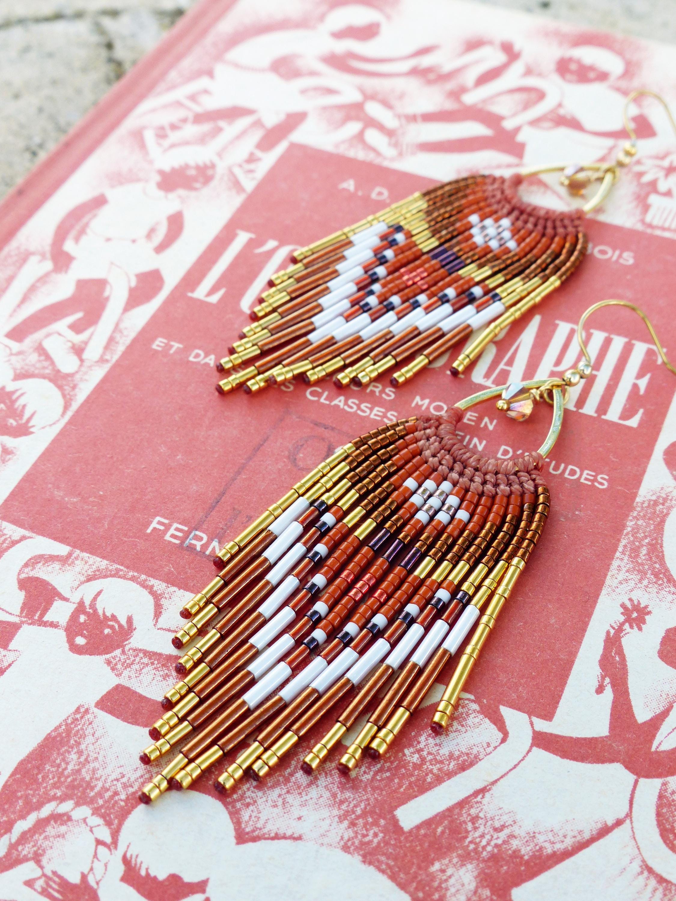 Peut inclure: Une paire de boucles d'oreilles en perles avec un motif de franges. Les boucles d'oreilles présentent un motif de perles blanches, rouges et dorées. Les boucles d'oreilles sont posées sur un fond rouge.
