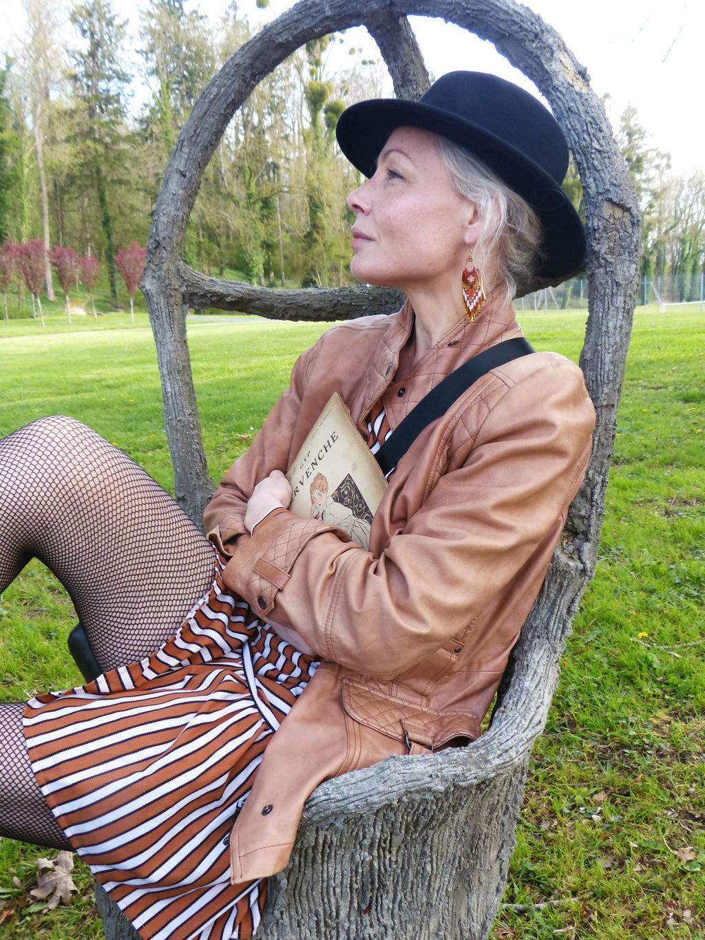 Peut inclure: Une personne portant un chapeau noir, une veste en cuir marron et une robe rayée est assise dans un fauteuil en tronc d'arbre. Elle tient un livre avec le titre "L'Aventure" visible. L'arrière-plan présente un champ vert et des arbres.