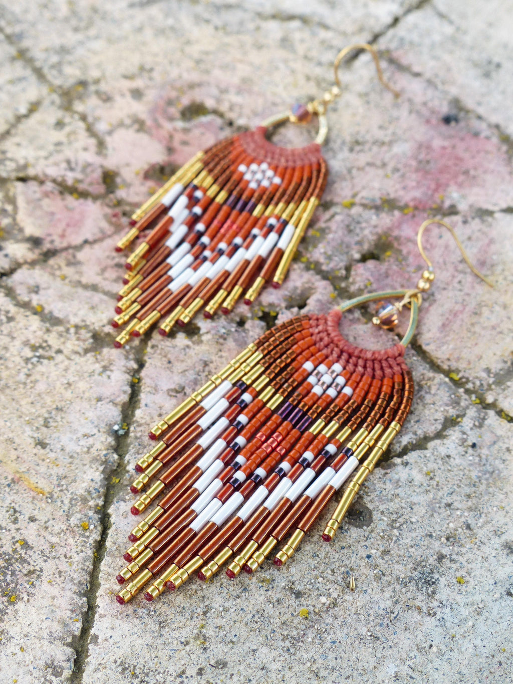 Peut inclure: Une paire de boucles d'oreilles faites à la main avec un design bohème. Les boucles d'oreilles présentent une combinaison de perles orange, blanches et dorées, disposées selon un motif géométrique. Les boucles d'oreilles sont de style frange.