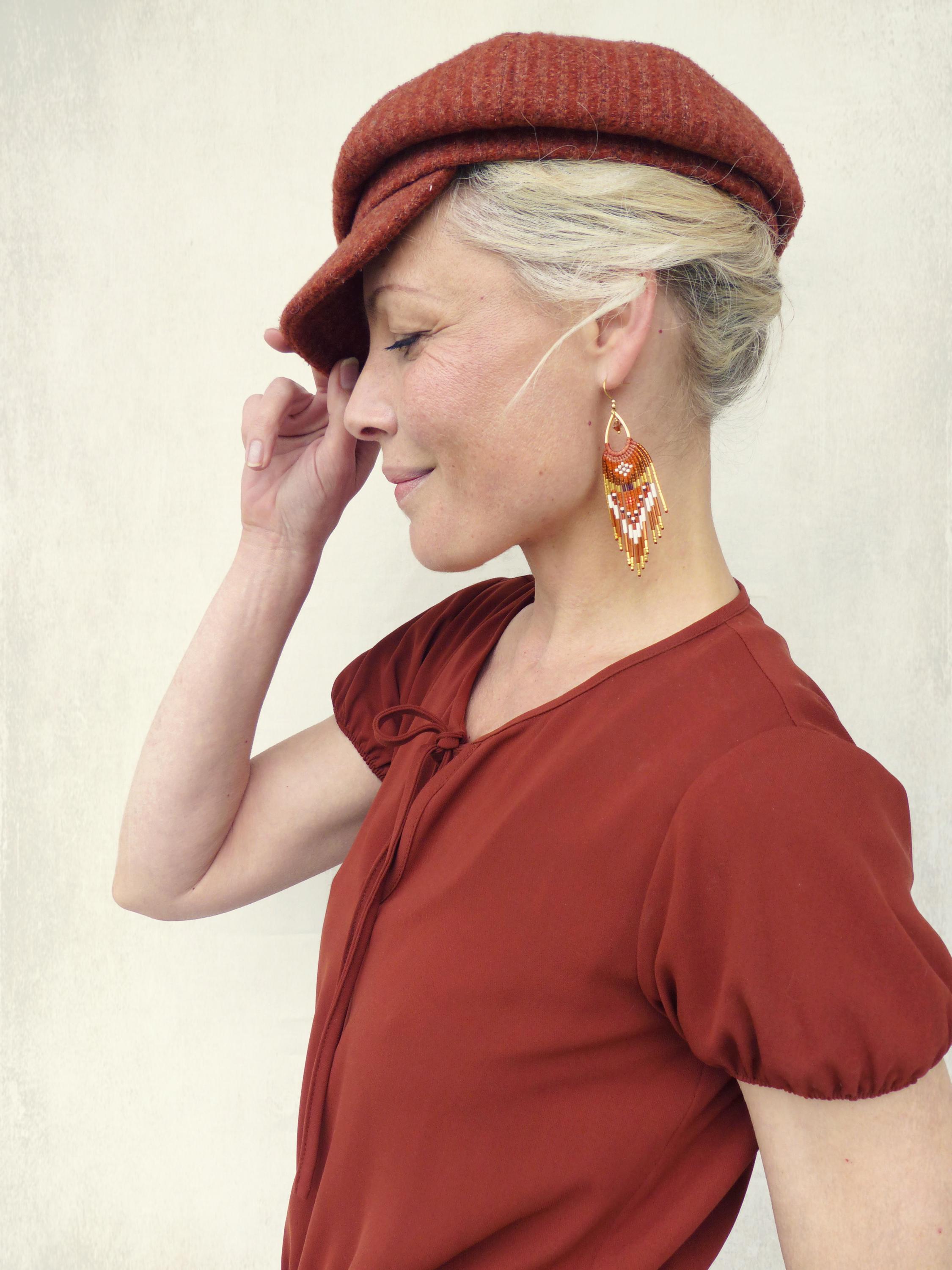 Peut inclure: Une femme porte une casquette de style gavroche couleur rouille et un haut à manches courtes assorti. Elle porte également de longues boucles d'oreilles pendantes avec des perles orange, blanches et dorées. La femme sourit et regarde vers le bas.