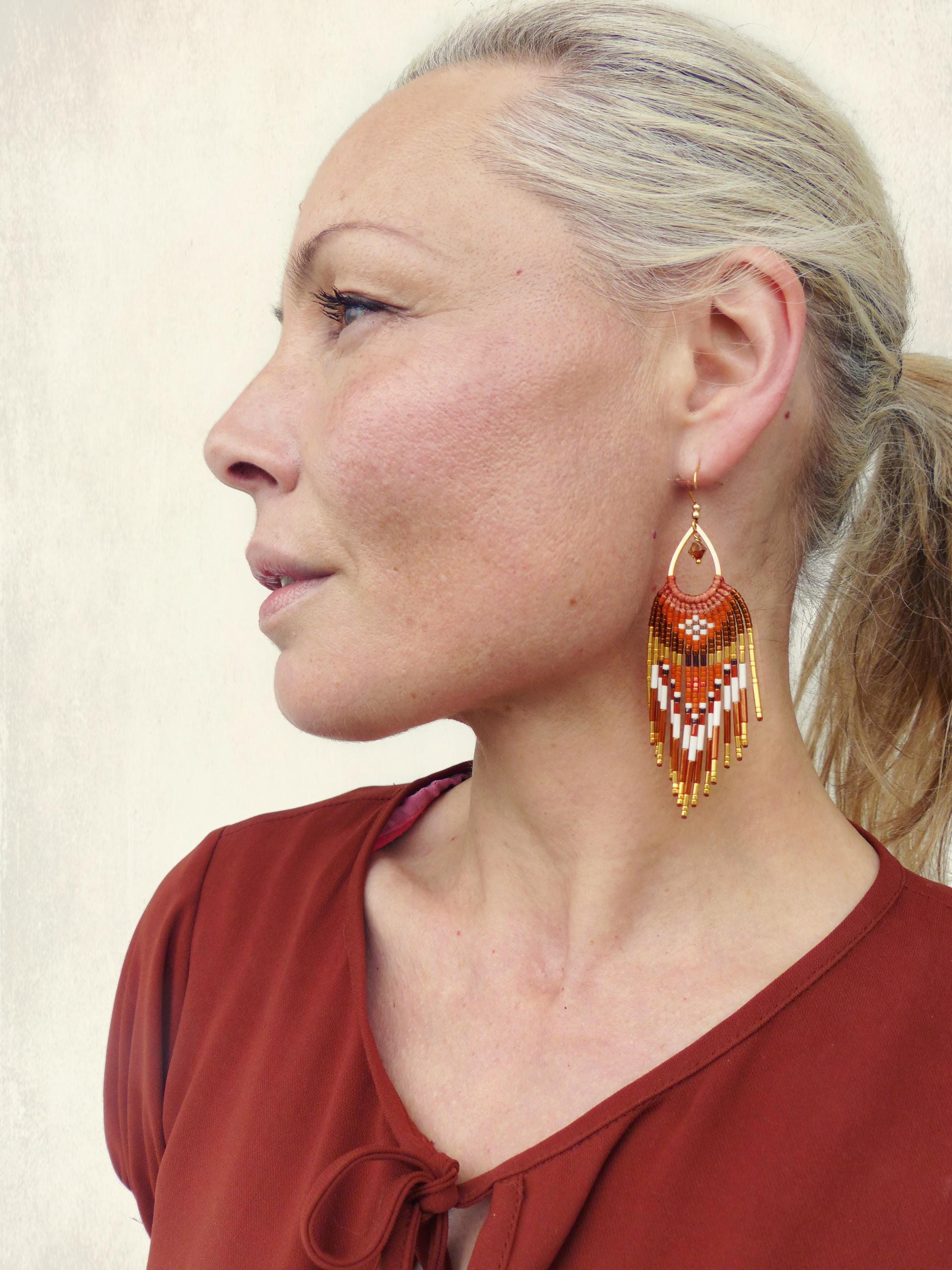 Peut inclure: Boucles d'oreilles en perles orange, blanches et dorées. Elles présentent un motif géométrique en forme de goutte d'eau et des accents métalliques dorés. Elles sont suspendues à un crochet doré. Un accessoire saisissant.