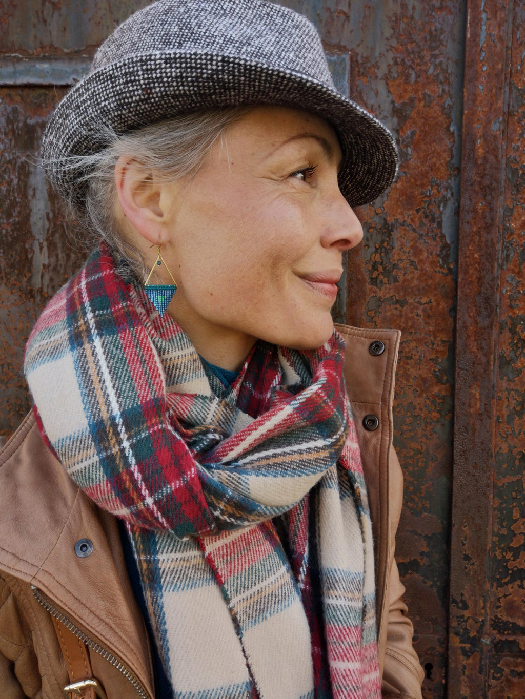 Peut inclure: Une femme portant un chapeau gavroche en tweed gris, une écharpe à carreaux rouge, verte et crème, et des boucles d'oreilles triangulaires dorées avec des perles bleues et turquoise. Elle porte également une veste en cuir marron.