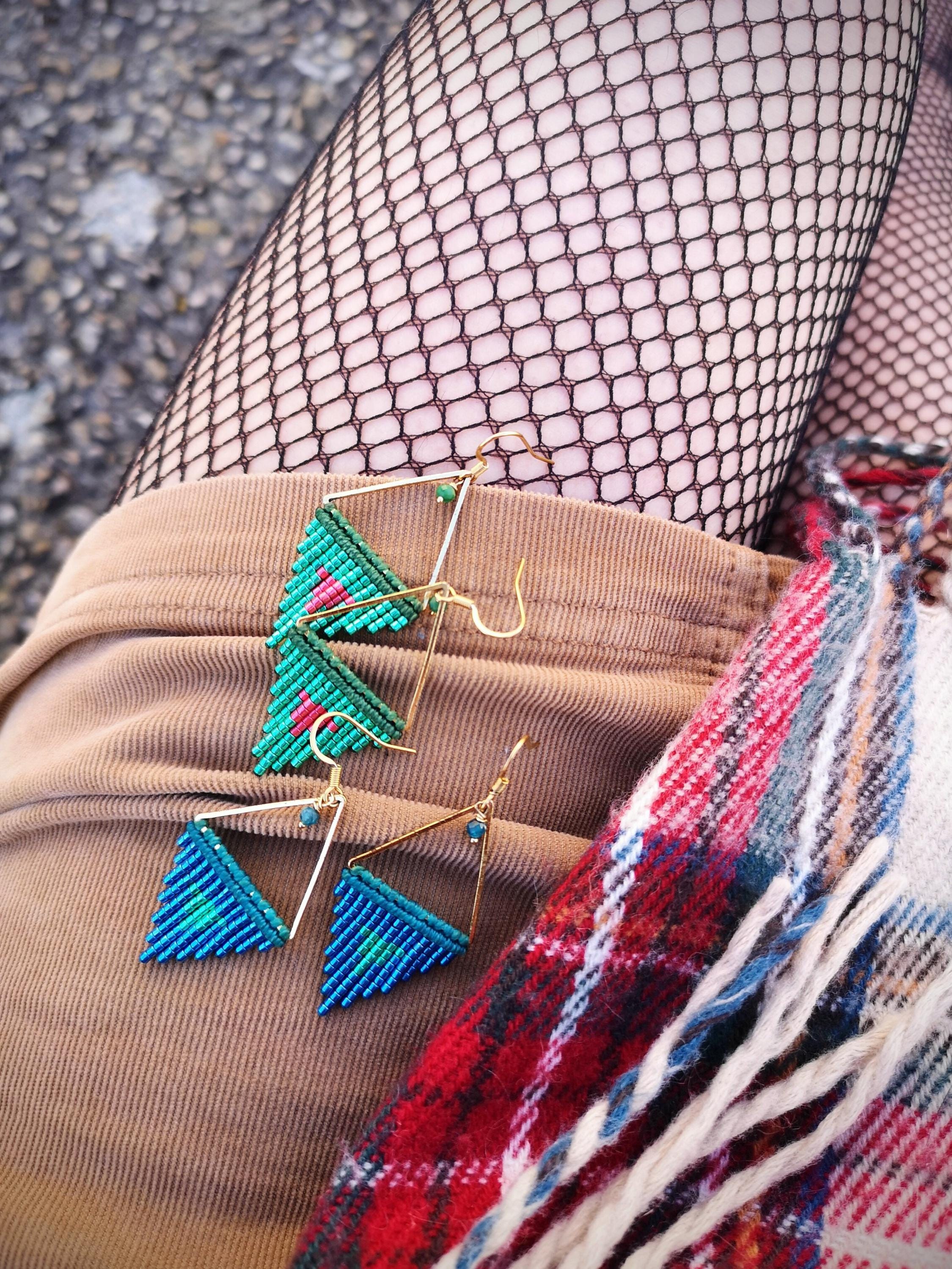 Peut inclure: Deux paires de boucles d'oreilles macramé faites à la main. Une paire est turquoise et bleue, l'autre est turquoise, rose et verte. Les deux paires ont des montures triangulaires en métal doré et de petites perles bleues. Les boucles d'oreilles sont présentées sur une surface en velours côtelé marron.