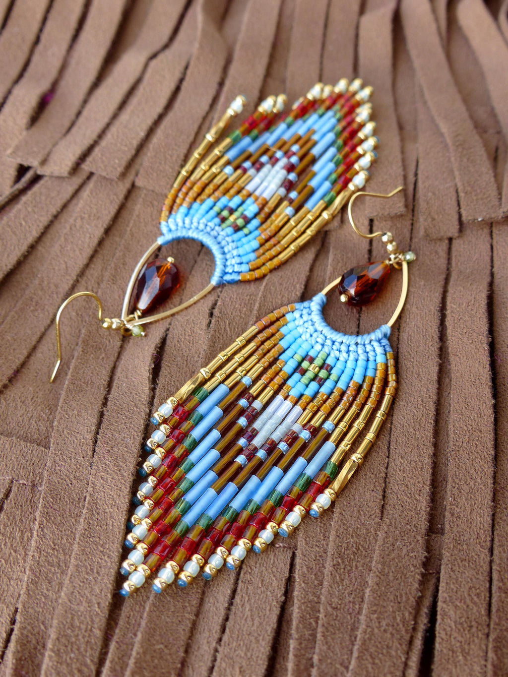 Peut inclure: Paire de boucles d'oreilles faites à la main avec un motif de plumes. Les boucles d'oreilles sont composées de perles bleues, rouges, blanches et dorées. Chaque boucle d'oreille a une perle ambrée en forme de goutte et des crochets d'oreilles dorés.