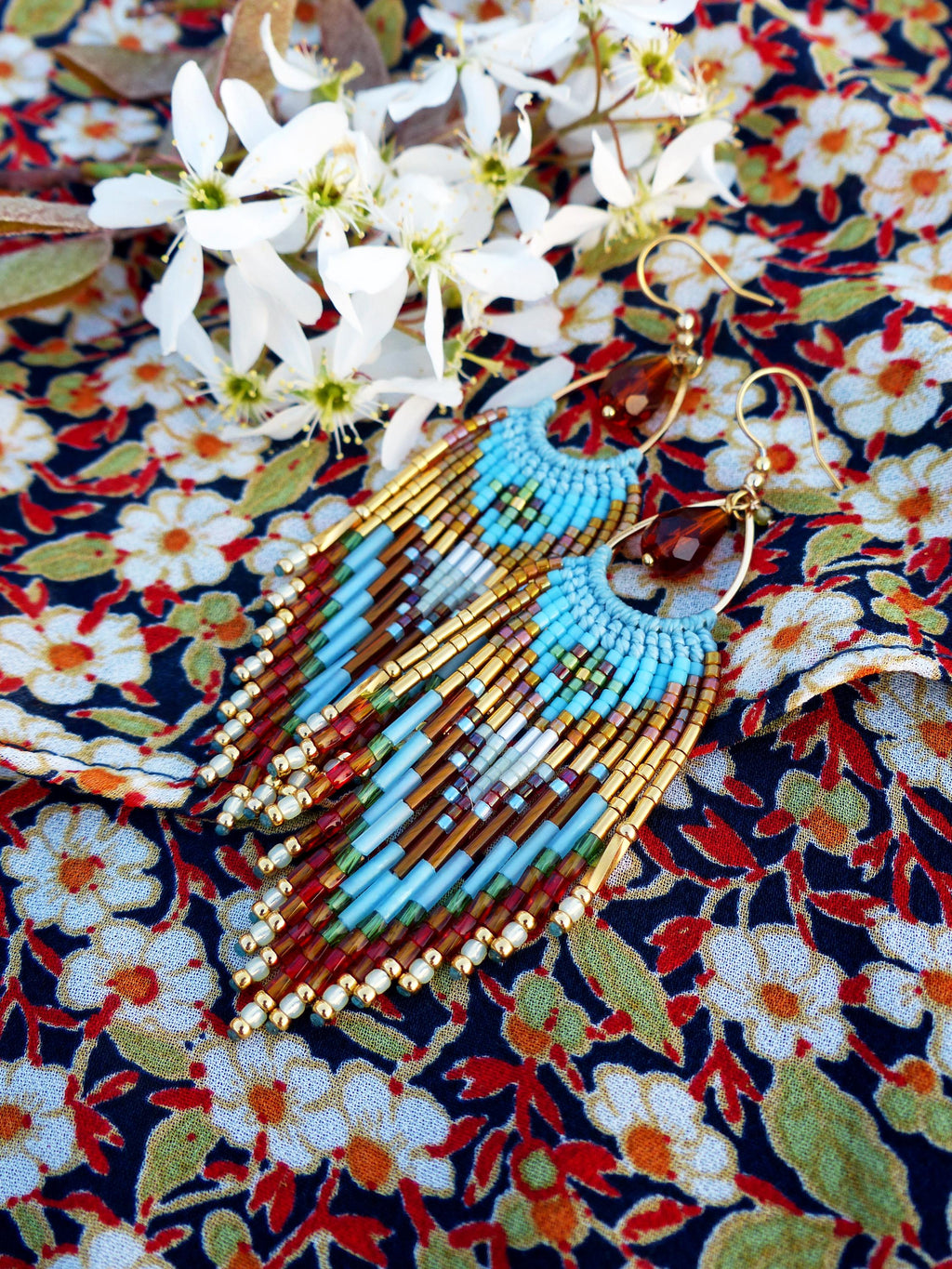 Peut inclure: Boucles d'oreilles faites à la main avec un design à franges. Les boucles d'oreilles présentent une combinaison de perles bleues, dorées, marron et rouges. Elles sont présentées sur un tissu à motifs floraux.