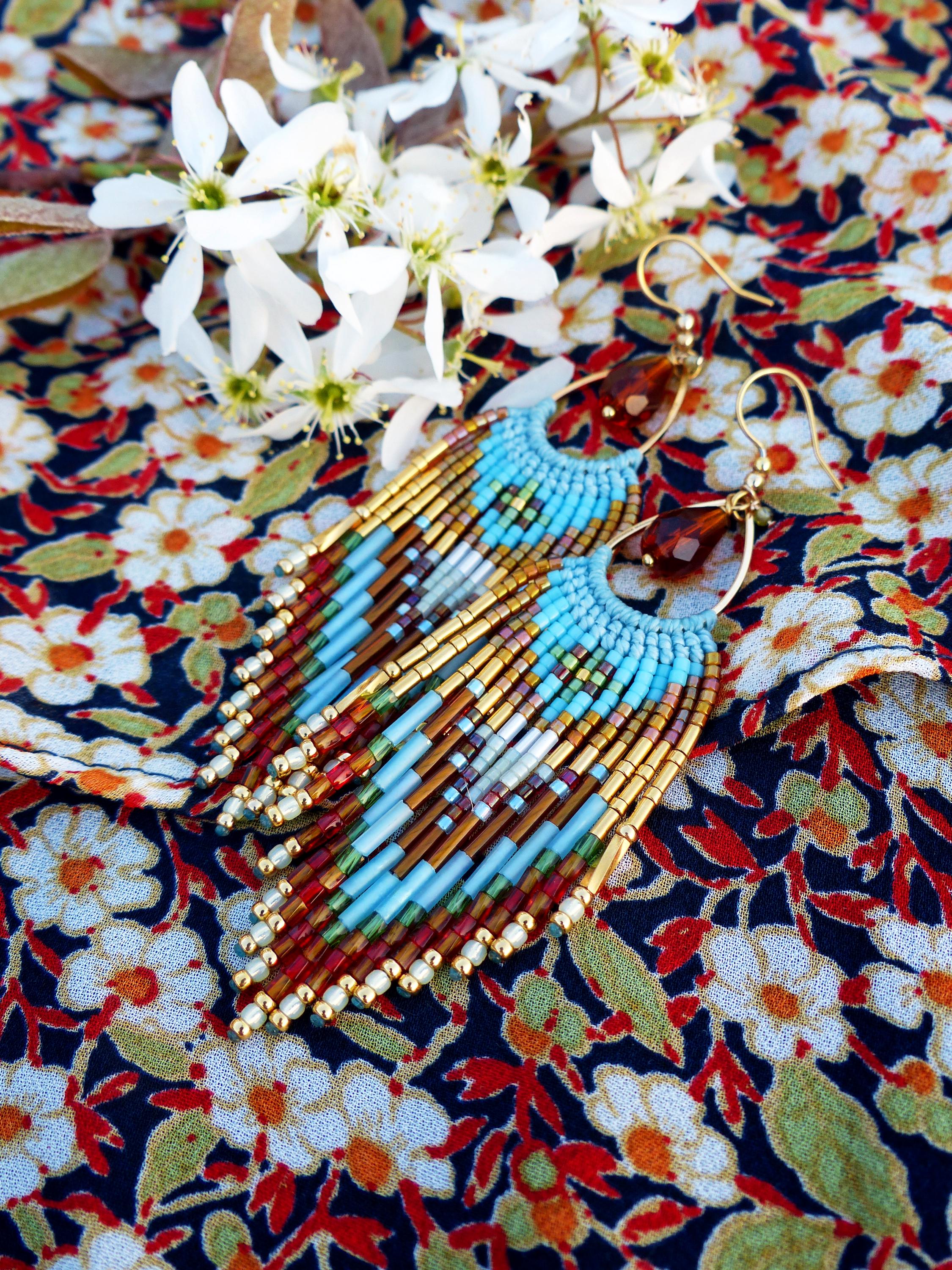 Peut inclure: Boucles d'oreilles faites à la main avec un design à franges. Les boucles d'oreilles présentent une combinaison de perles bleues, dorées, marron et rouges. Elles sont présentées sur un tissu à motifs floraux.