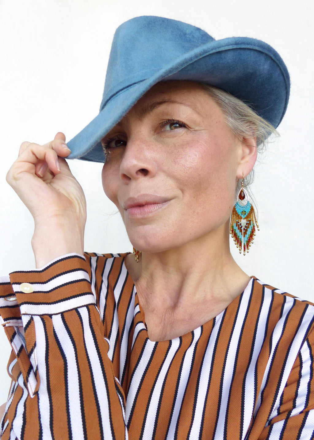 Peut inclure: Une femme porte un chapeau en feutre bleu clair et des boucles d'oreilles perlées. Les boucles d'oreilles ont une forme de goutte d'eau avec des accents rouges, turquoise et dorés. Elle porte une chemise rayée marron et blanche.