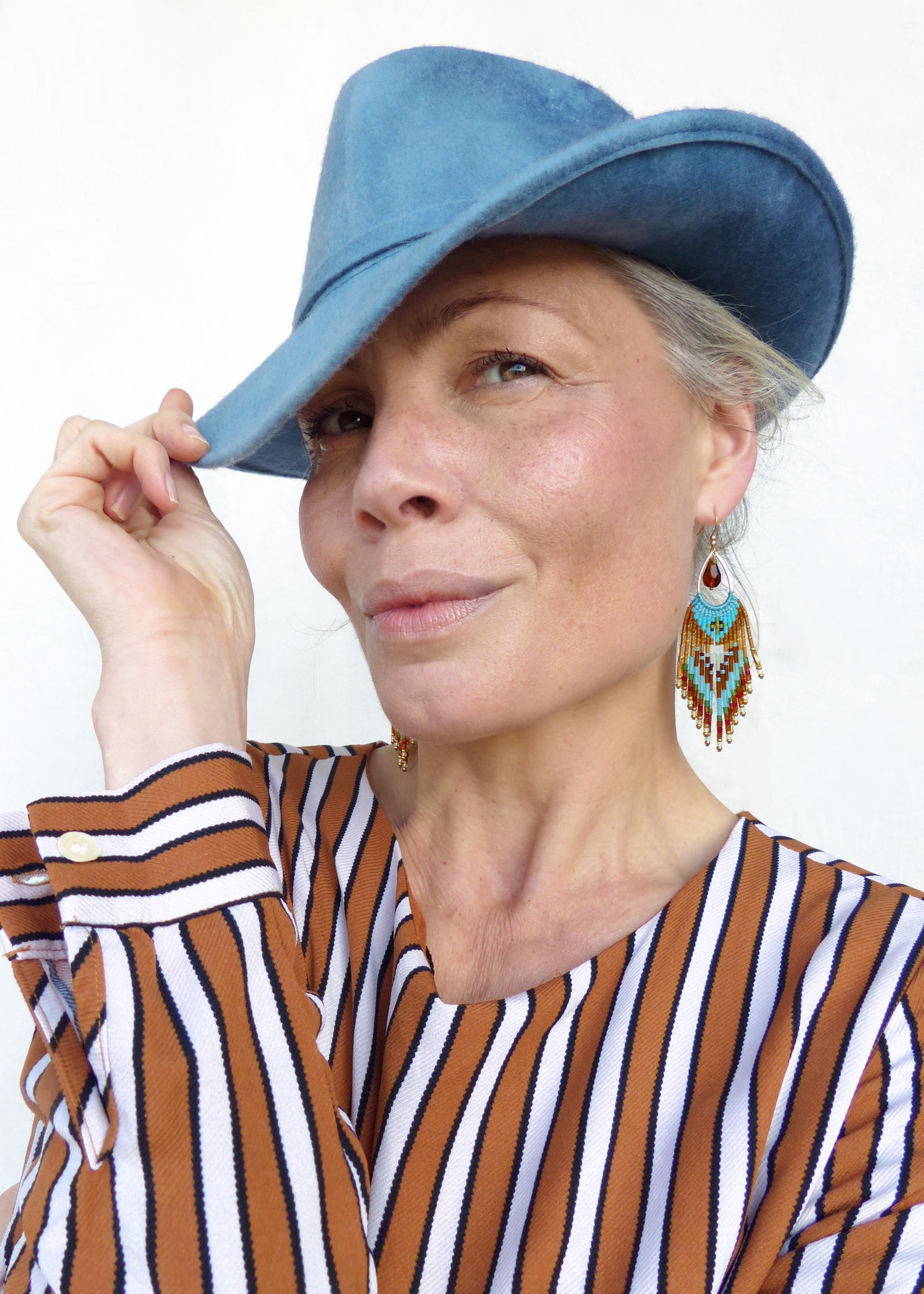 Peut inclure: Une femme porte un chapeau en feutre bleu clair et des boucles d'oreilles perlées. Les boucles d'oreilles ont une forme de goutte d'eau avec des accents rouges, turquoise et dorés. Elle porte une chemise rayée marron et blanche.