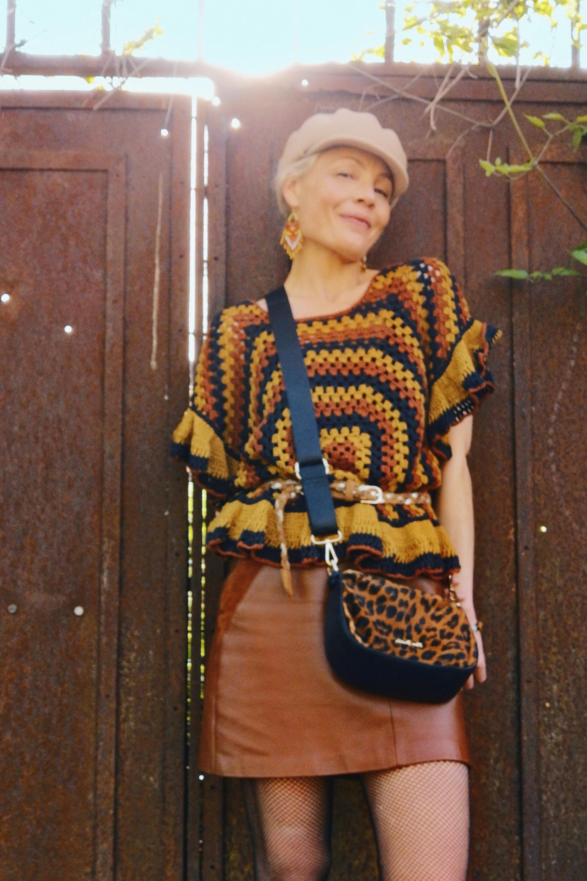 Peut inclure: Une femme porte un haut en crochet fait à la main dans les tons marron, jaune et bleu marine, associé à une jupe en cuir marron et un sac bandoulière à imprimé léopard. Elle porte également une casquette beige et des boucles d'oreilles.