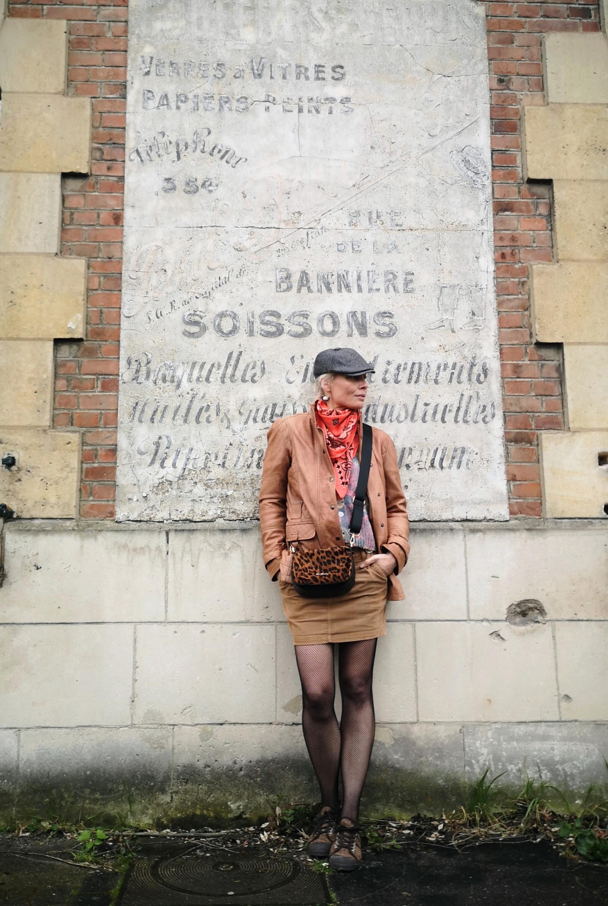 Peut inclure: Une personne porte une veste en cuir marron, une jupe beige et un sac à bandoulière à imprimé léopard. La personne se tient devant un mur de pierre vieilli avec du texte français délavé.