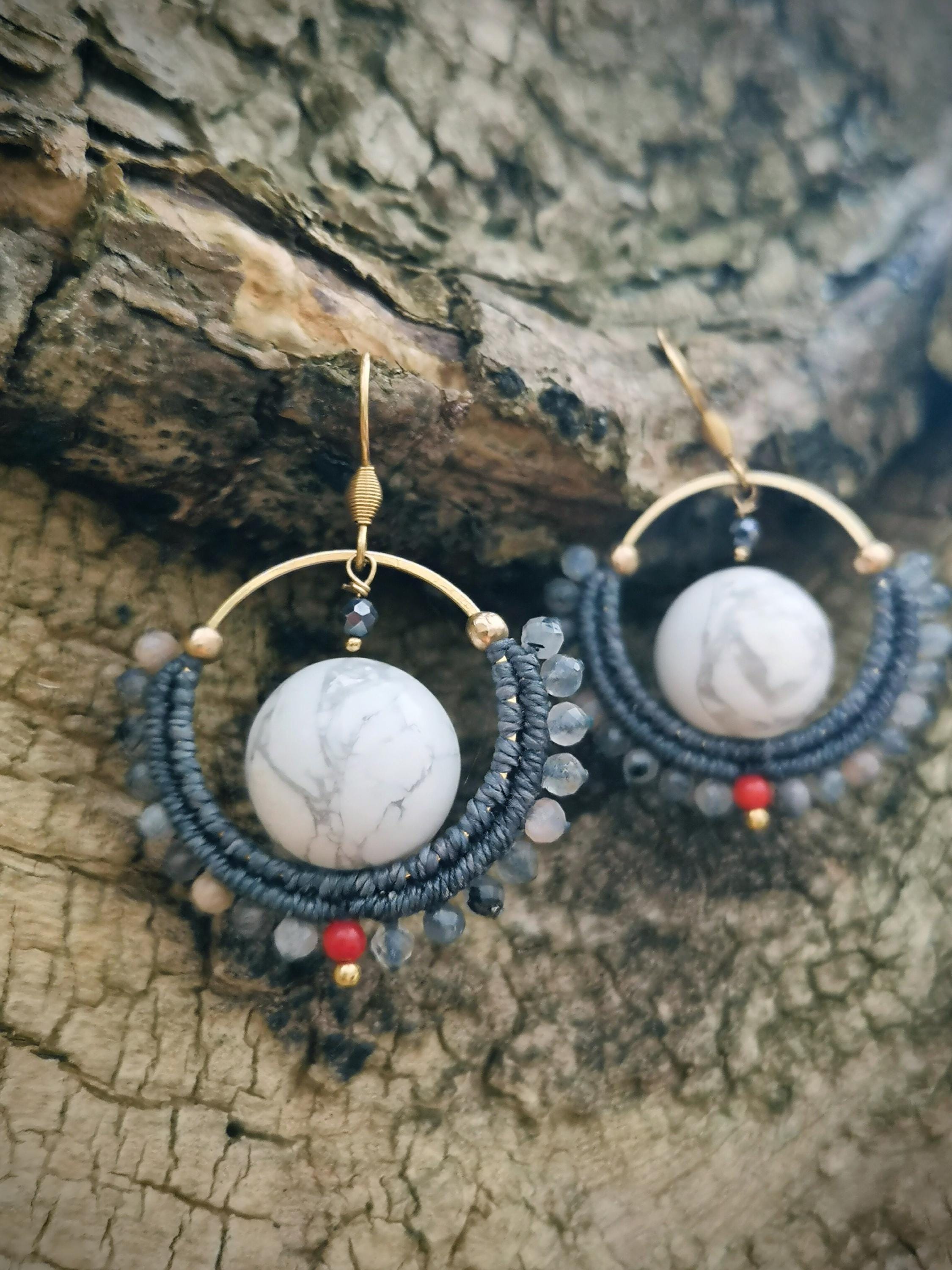 Peut inclure: Boucles d'oreilles faites à la main avec une sphère en marbre blanc suspendue dans un croissant en macramé gris foncé. Les boucles d'oreilles ont des crochets et des accents dorés, avec de petites perles rouges et grises. Fond texturé brun.