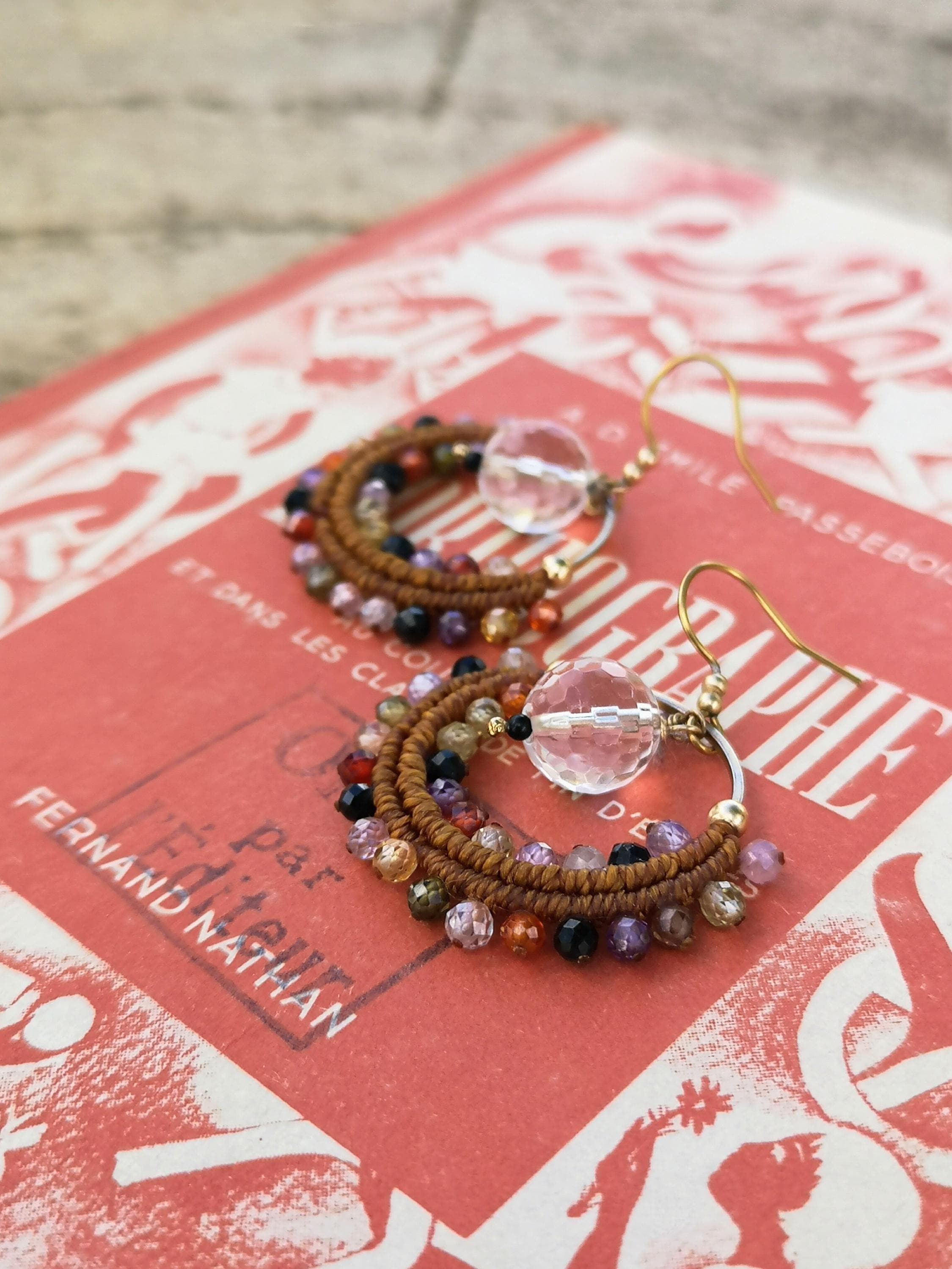 Peut inclure: Boucles d'oreilles créoles faites à la main avec une perle de cristal transparente et un design circulaire. Les créoles sont ornées d'une variété de perles colorées dans les tons marron, violet, orange et noir, créant un style bohème.