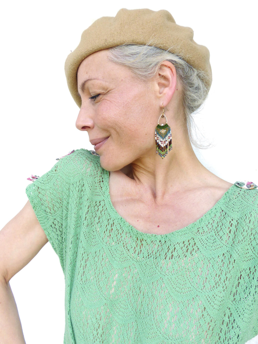 Peut inclure: Une femme porte un béret beige et un haut vert clair avec un motif en dentelle. Elle porte des boucles d'oreilles dorées avec des franges de perles vertes, marron et blanches. Le haut a un col rond et des manches courtes.