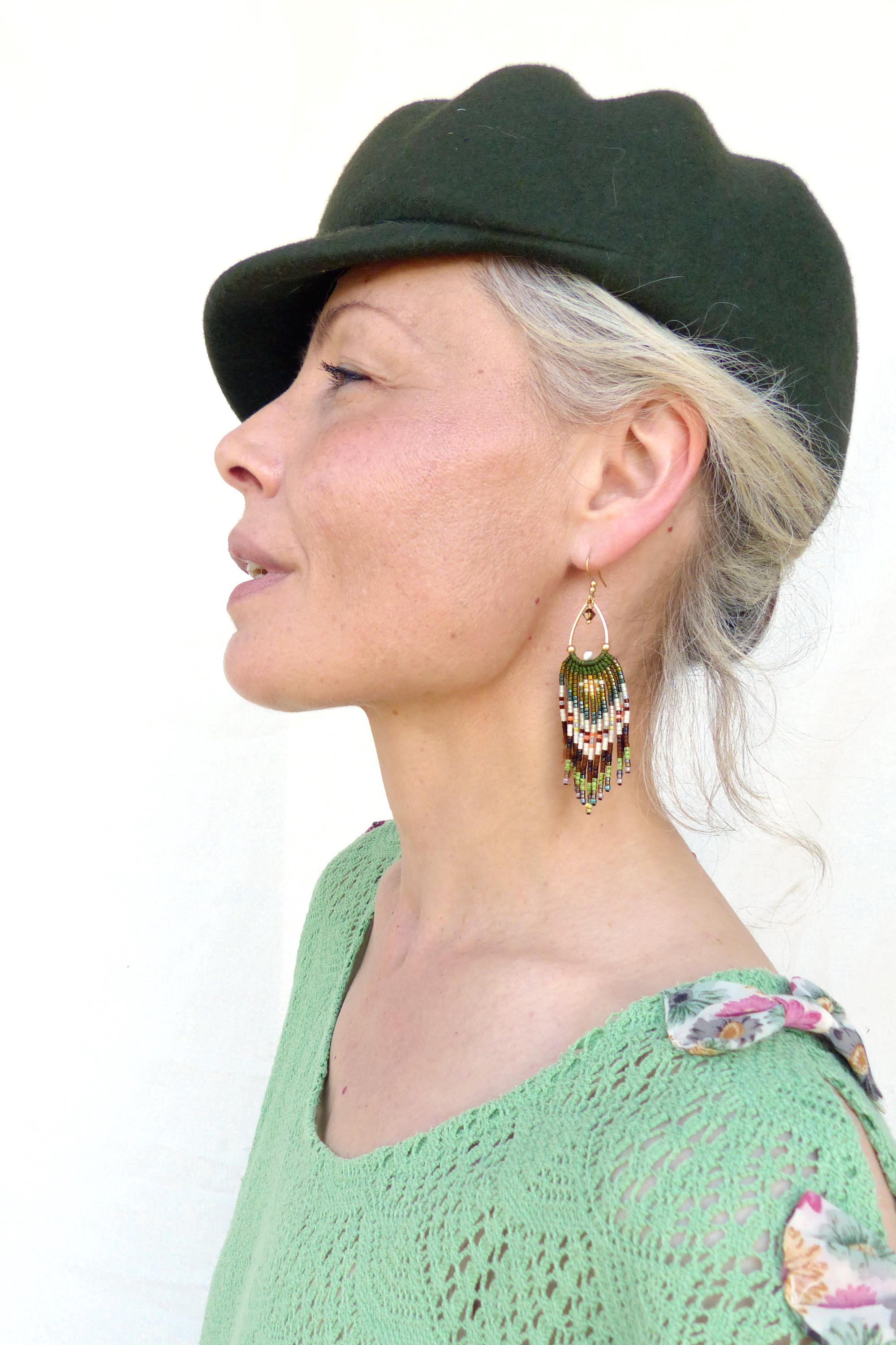 Peut inclure: Vue de profil d'une personne portant une casquette vert foncé et un haut vert clair. La personne porte des boucles d'oreilles dorées à franges de perles vertes, rouges et blanches.