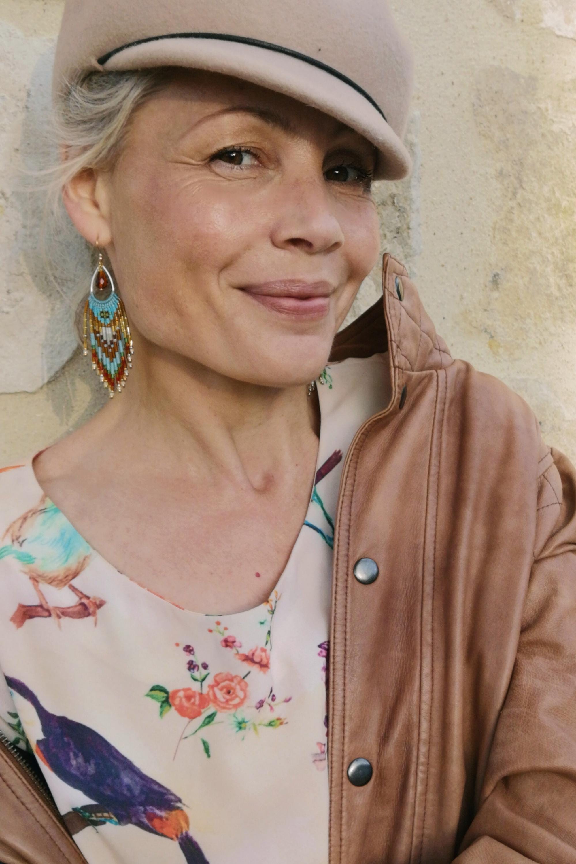 Peut inclure: Une femme porte une casquette beige et une veste en cuir marron. Elle porte également des boucles d'oreilles en perles avec des accents turquoise, rouges et marron. Le chemisier à imprimé floral présente des oiseaux et des fleurs.