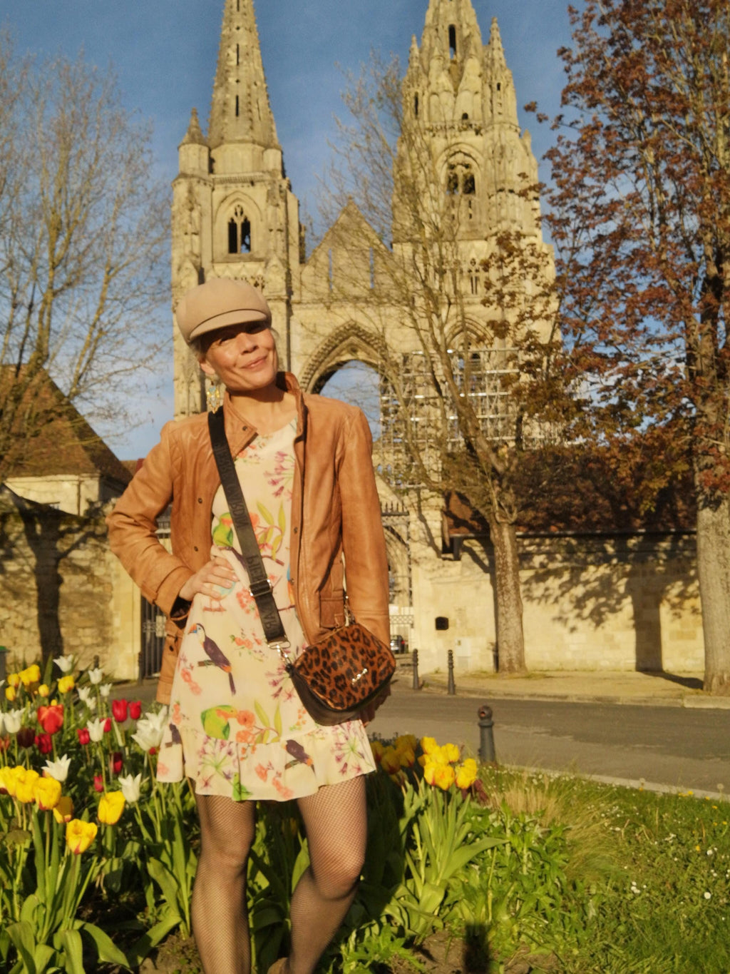 Peut inclure: Une personne porte une casquette beige, une veste en cuir marron et une robe à imprimé floral avec des illustrations d'oiseaux. Elle porte un petit sac à bandoulière à imprimé léopard. L'arrière-plan présente un bâtiment historique en pierre et un parterre de fleurs avec des tulipes colorées.