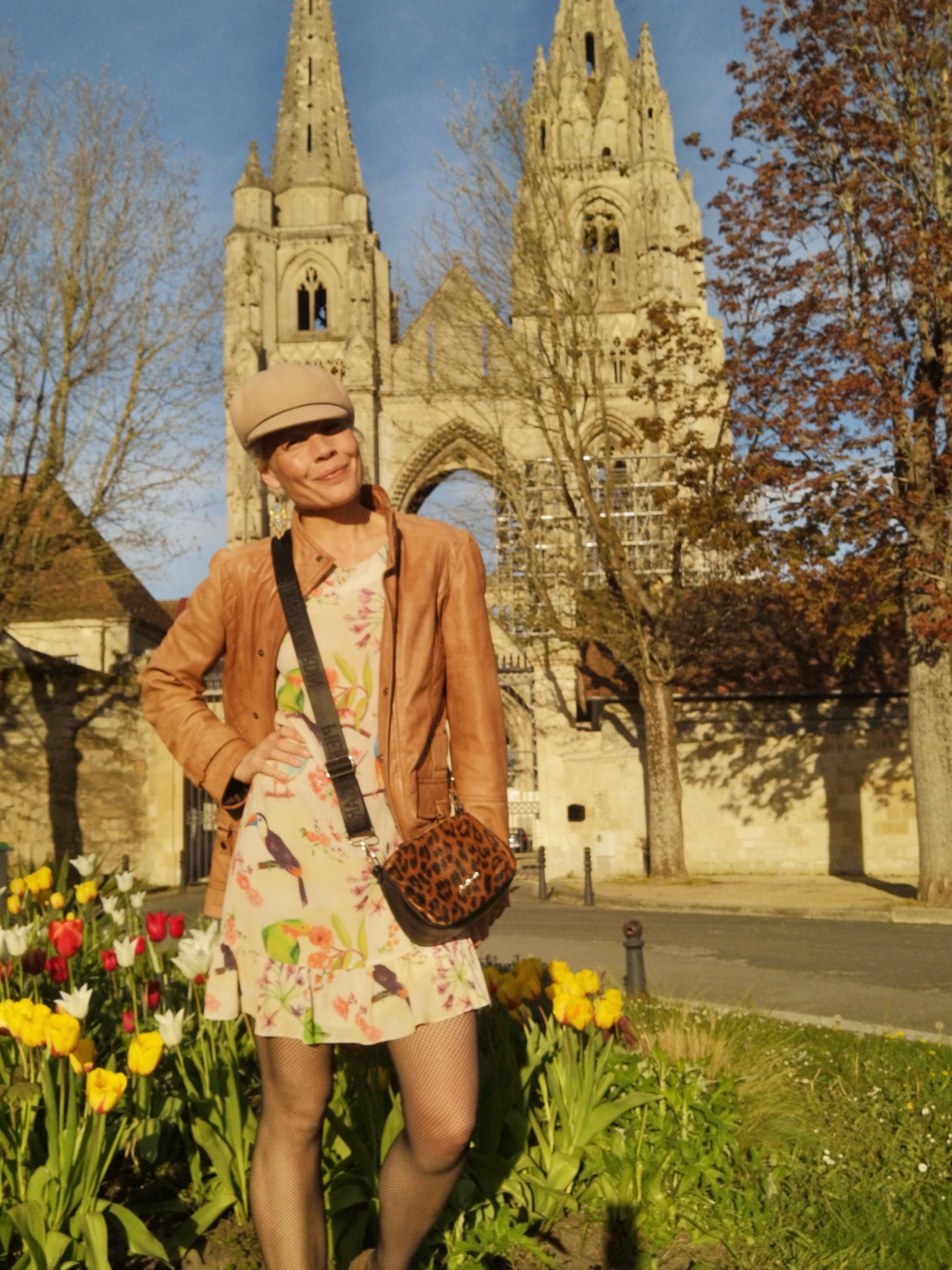 Peut inclure: Une personne porte une casquette beige, une veste en cuir marron et une robe à imprimé floral avec des illustrations d'oiseaux. Elle porte un petit sac à bandoulière à imprimé léopard. L'arrière-plan présente un bâtiment historique en pierre et un parterre de fleurs avec des tulipes colorées.