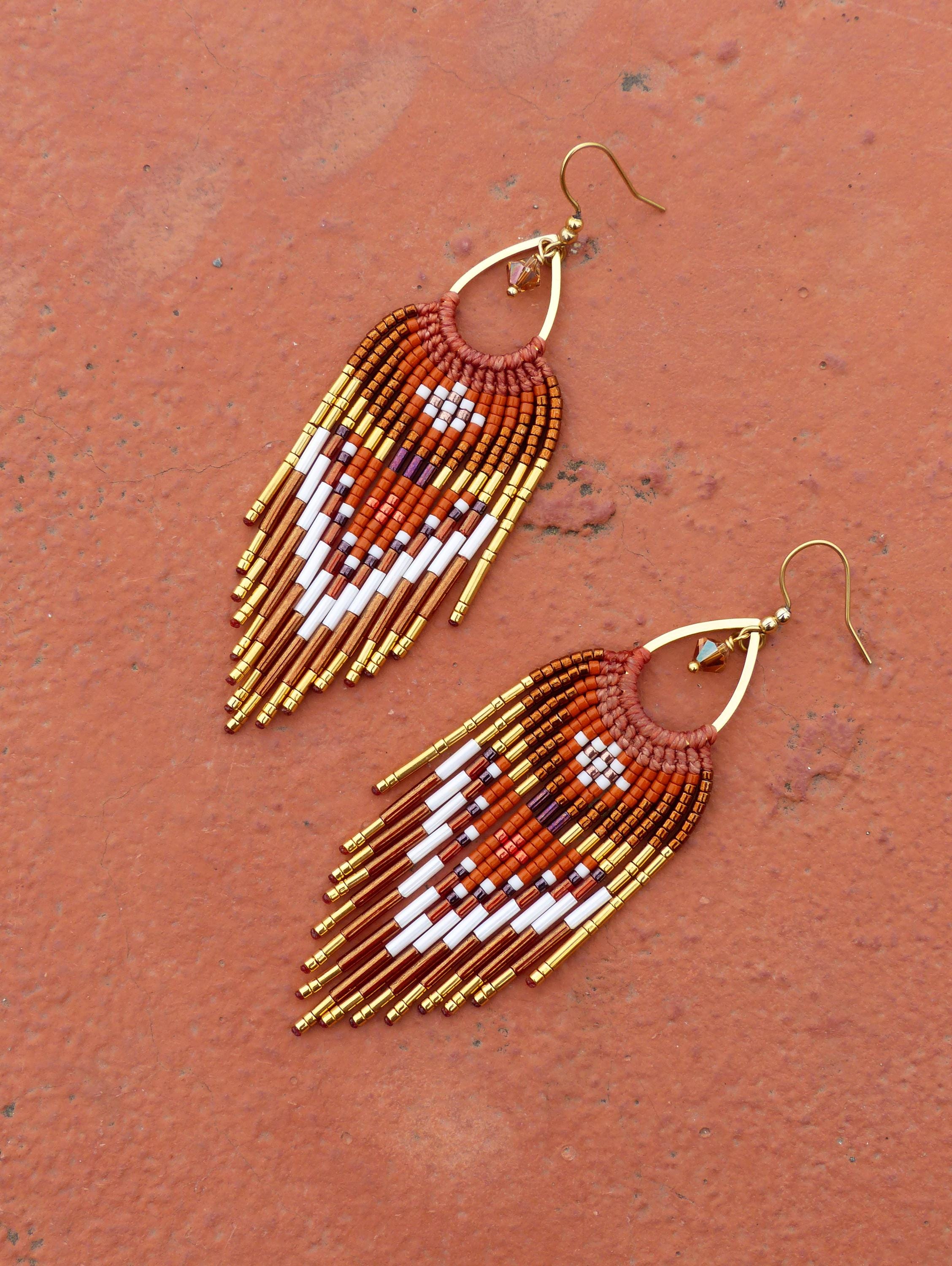 Peut inclure: Une paire de boucles d'oreilles perlées faites à la main. Les boucles d'oreilles présentent une forme de goutte d'eau avec un crochet doré et un motif de perles dans les tons orange, blanc et or. Les boucles d'oreilles ont une frange de perles dorées.
