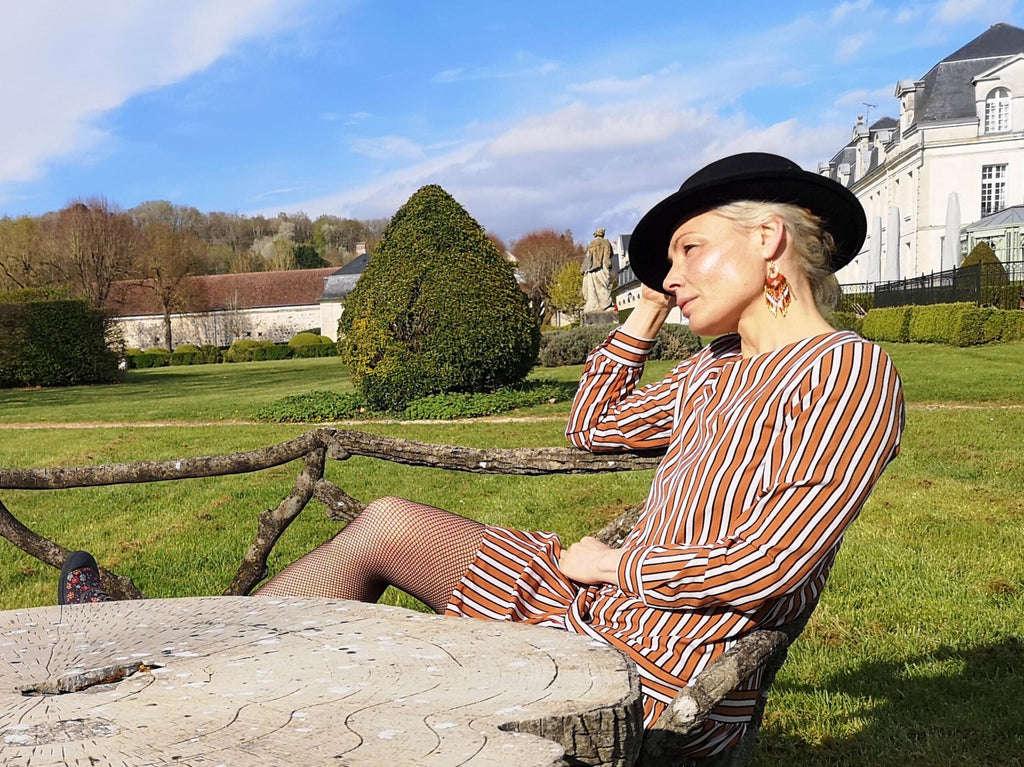 Peut inclure: Robe à rayures marron et blanches et chapeau noir. La robe présente des rayures verticales et des manches longues. Le chapeau est noir et rond. La personne est assise sur un banc en bois dans un cadre de parc.