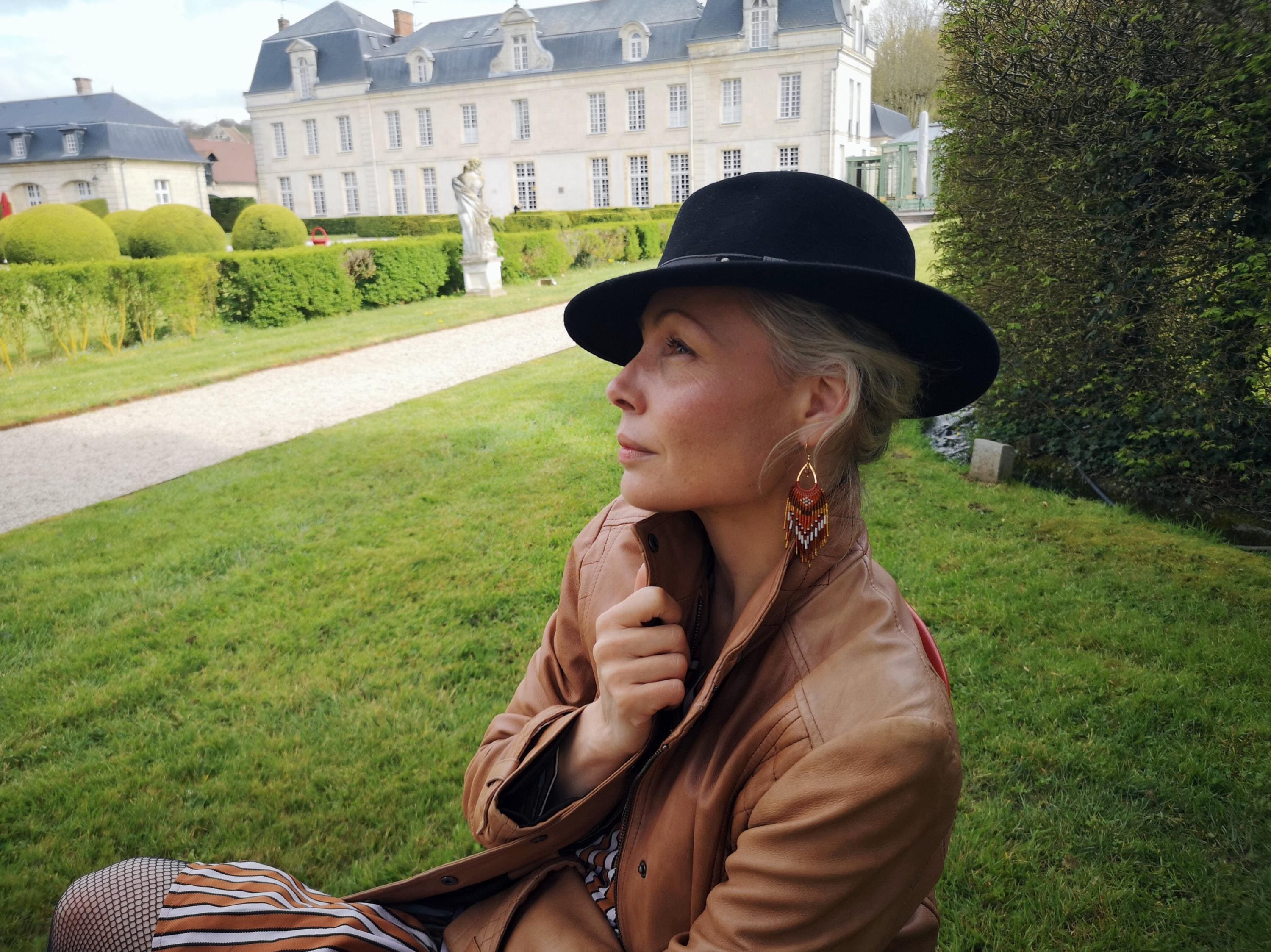 Peut inclure: Une femme portant un chapeau fedora noir et une veste en cuir marron, assise sur de l'herbe. La femme porte des boucles d'oreilles. Un grand bâtiment est en arrière-plan.