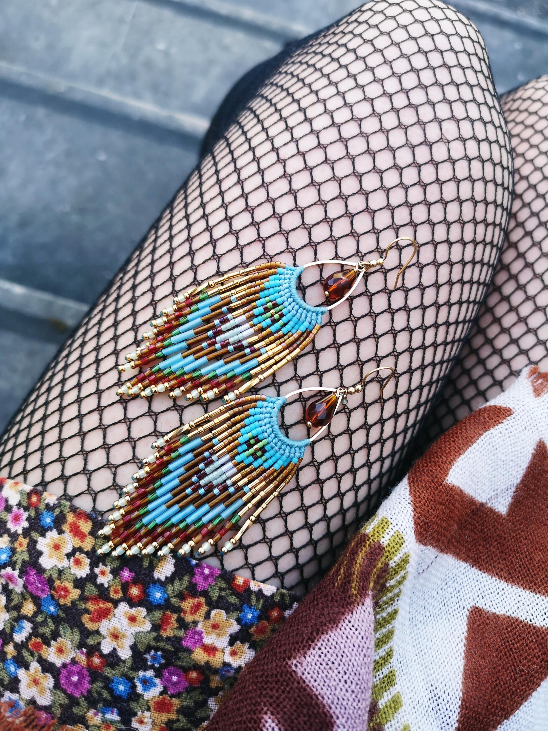 Peut inclure: Une paire de boucles d'oreilles faites à la main avec un design unique. Les boucles d'oreilles présentent une combinaison de perles bleues, marron et dorées, disposées en franges. Elles sont suspendues à des crochets dorés, créant un accessoire élégant.