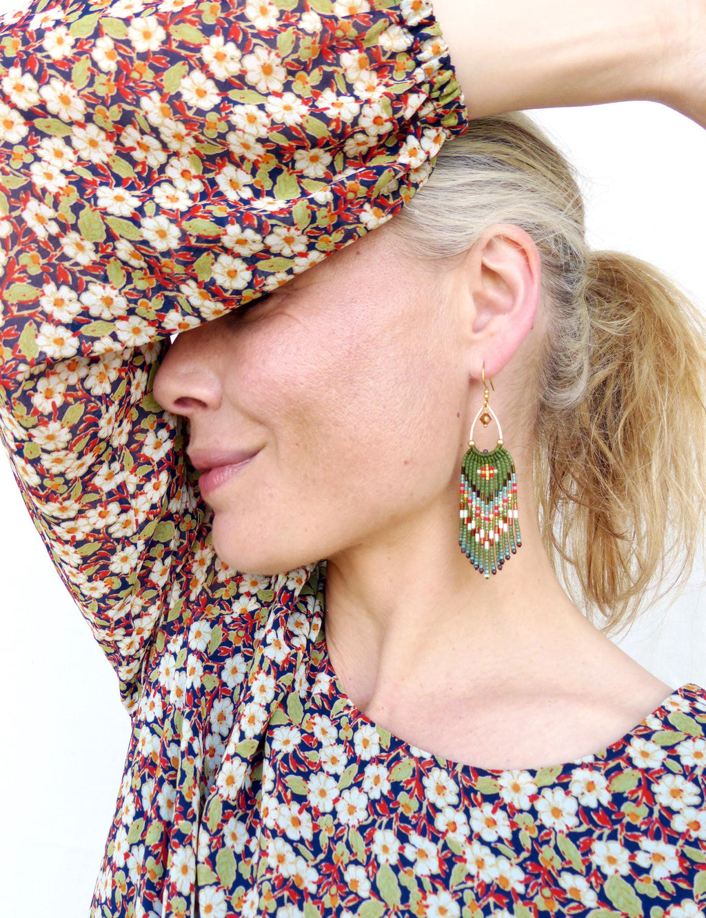 Peut inclure: Une femme porte des boucles d'oreilles en perles avec un dessus doré. Les boucles d'oreilles présentent un motif vert, rouge et blanc. La femme porte un haut à imprimé floral avec des fleurs blanches, des feuilles vertes et un fond bleu marine.