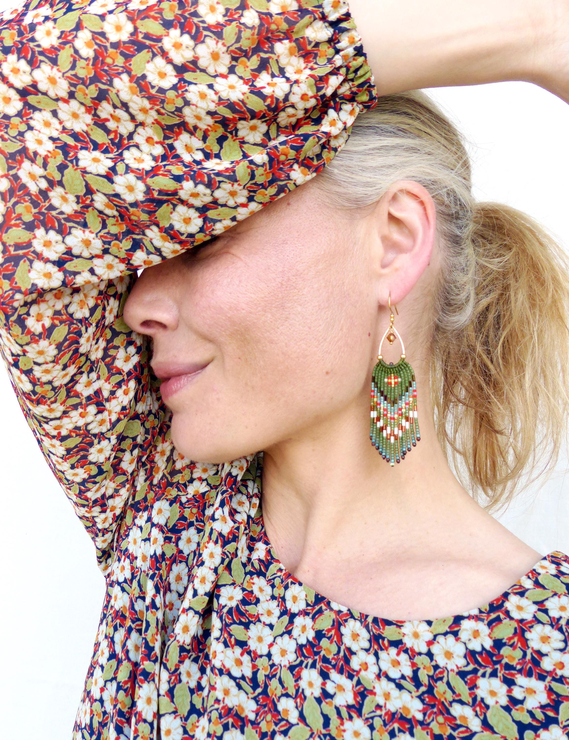 Peut inclure: Une femme porte des boucles d'oreilles en perles avec un dessus doré. Les boucles d'oreilles présentent un motif vert, rouge et blanc. La femme porte un haut à imprimé floral avec des fleurs blanches, des feuilles vertes et un fond bleu marine.