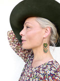Peut inclure: Une femme porte un chapeau vert foncé et un haut à imprimé floral. La femme porte des boucles d'oreilles en perles avec un motif vert, orange et blanc. Les boucles d'oreilles sont suspendues à des crochets dorés.