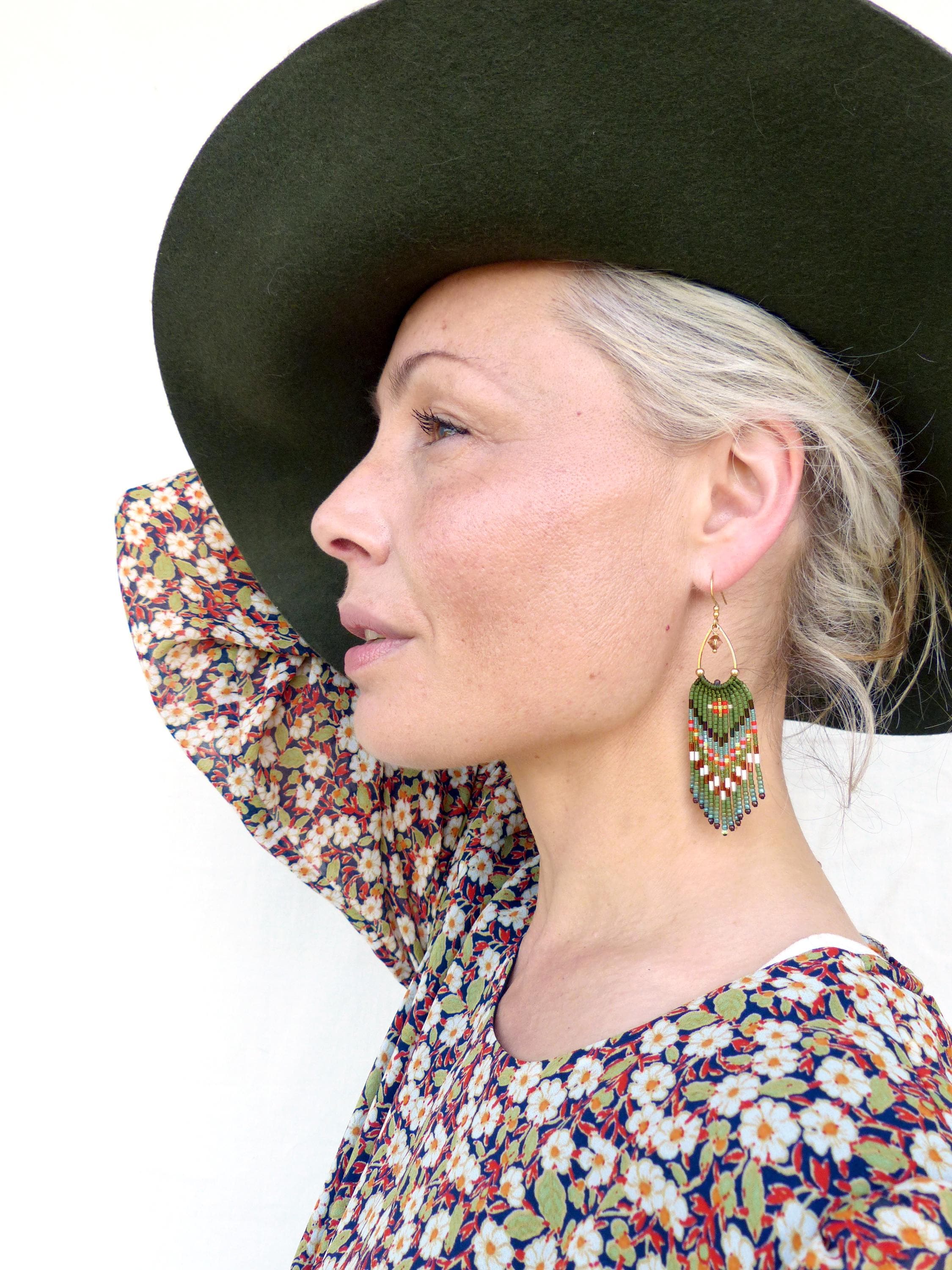 Peut inclure: Une femme porte un chapeau vert foncé et un haut à imprimé floral. La femme porte des boucles d'oreilles en perles avec un motif vert, orange et blanc. Les boucles d'oreilles sont suspendues à des crochets dorés.
