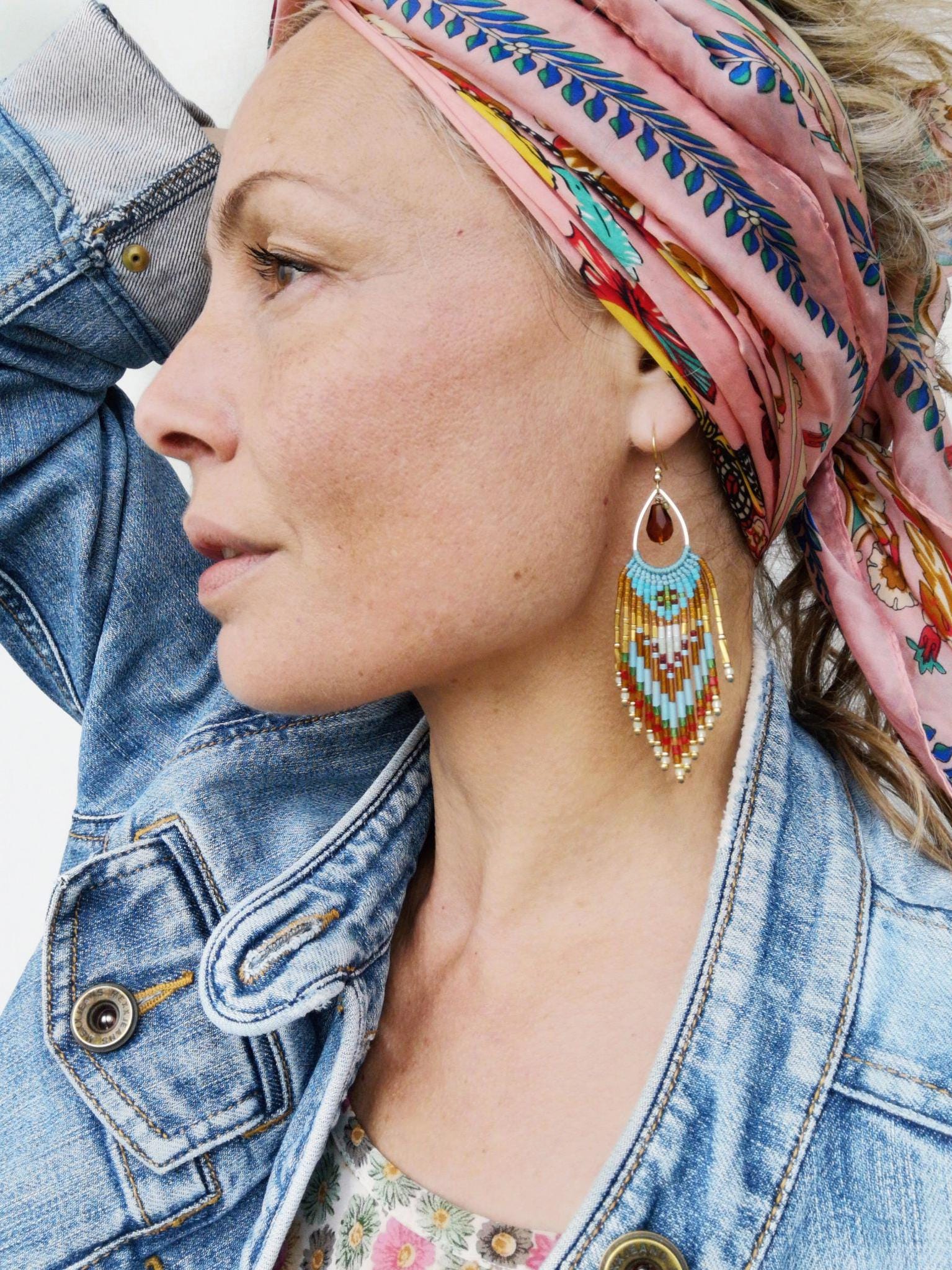 Peut inclure: Boucles d'oreilles dorées en forme de goutte d'eau avec une frange de perles turquoise, orange, blanche et dorée. Les boucles d'oreilles ont une petite pierre rouge foncé en haut. La personne porte un foulard rose clair avec un motif coloré et une veste en jean.
