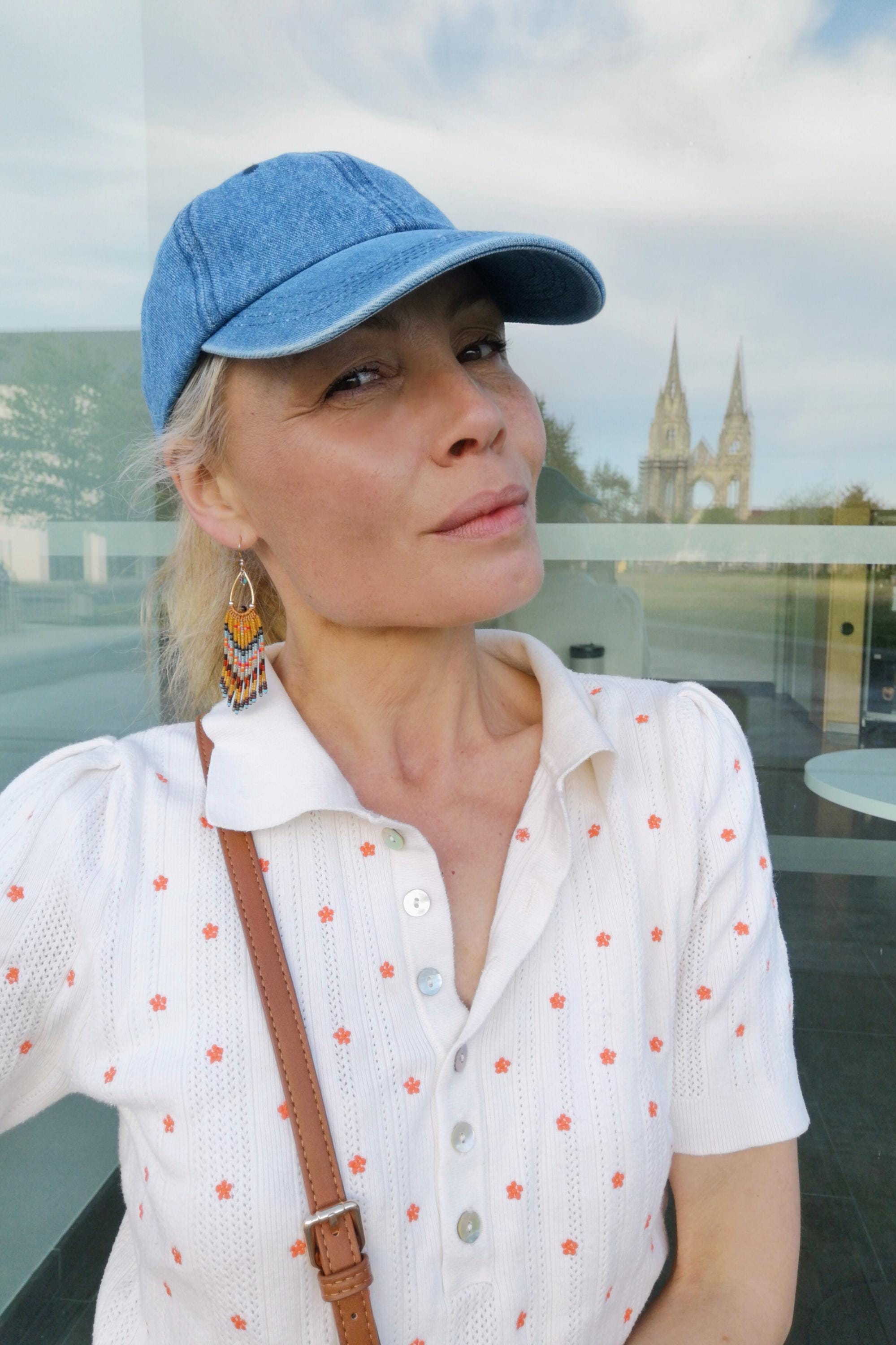 Peut inclure: Casquette de baseball en jean bleu clair. La personne porte une chemise blanche à col avec de petits motifs de fleurs orange. La personne porte également des boucles d'oreilles en perles de couleurs orange, jaune et bleu.