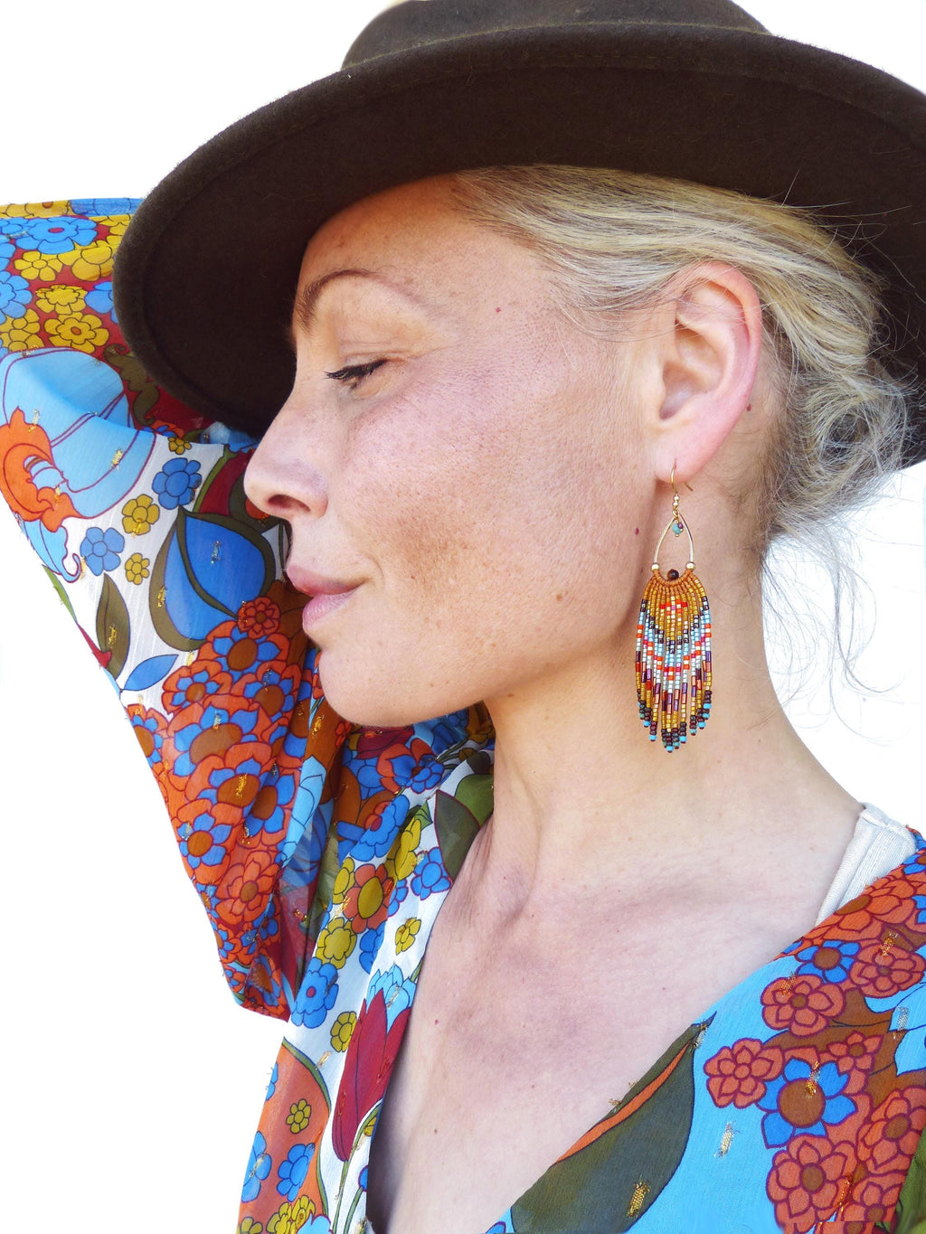 Peut inclure: Une femme porte un chapeau marron et une robe à motifs floraux colorés. L'attention se porte sur une paire de boucles d'oreilles en perles en forme de goutte d'eau, avec des perles orange, rouges, bleues et marron.