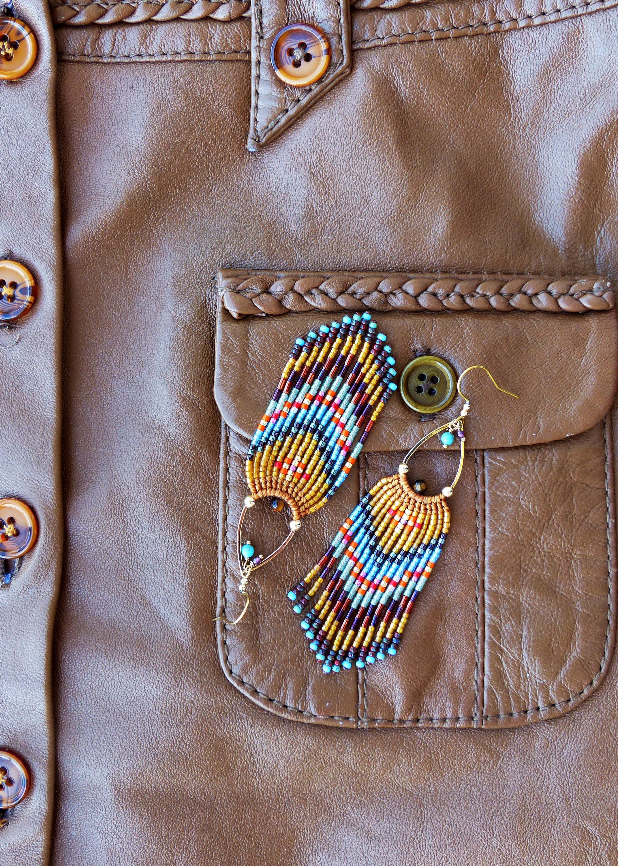 Peut inclure: Boucles d'oreilles à franges perlées. Les boucles d'oreilles présentent un motif de perles bleues, jaunes, oranges et marron. Elles sont posées sur une surface en cuir marron, avec un bouton et une poche visibles.