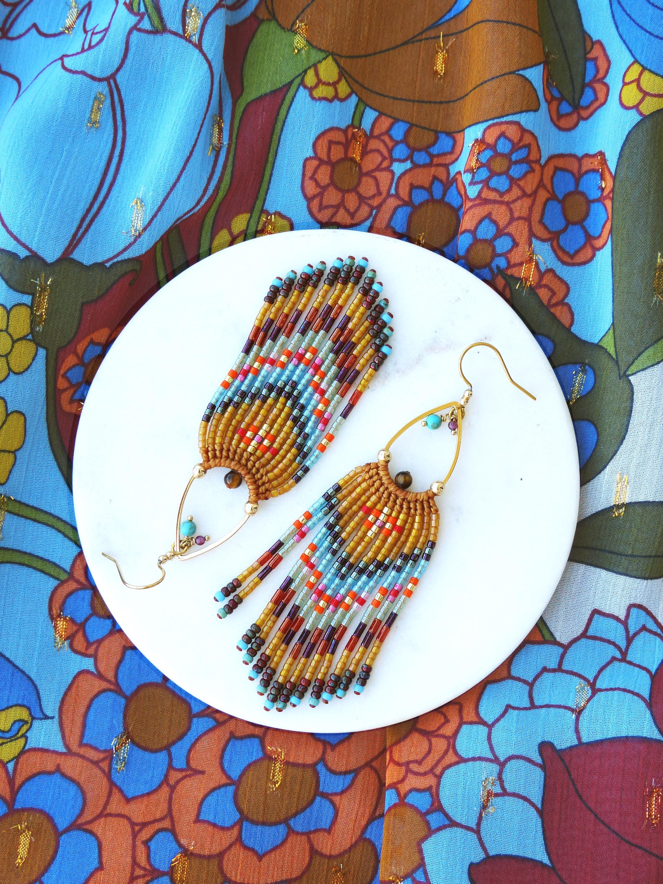 Peut inclure: Une paire de boucles d'oreilles en perles faites à la main avec un design à franges. Les boucles d'oreilles présentent une base dorée avec un motif de perles orange, bleues et rouges. Elles sont présentées sur une assiette blanche sur un fond floral coloré.