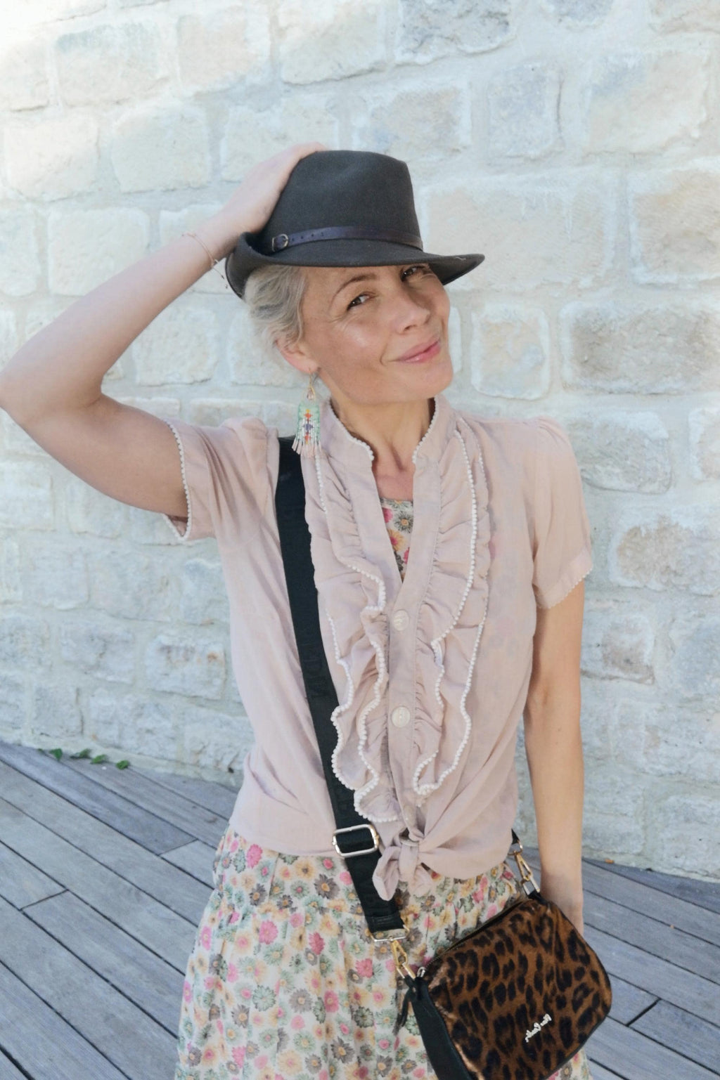Peut inclure: Une femme porte un chapeau fedora gris foncé avec une bande en cuir marron. Elle porte un chemisier rose clair avec des volants et une jupe à fleurs. Un sac bandoulière noir avec un imprimé léopard est visible.