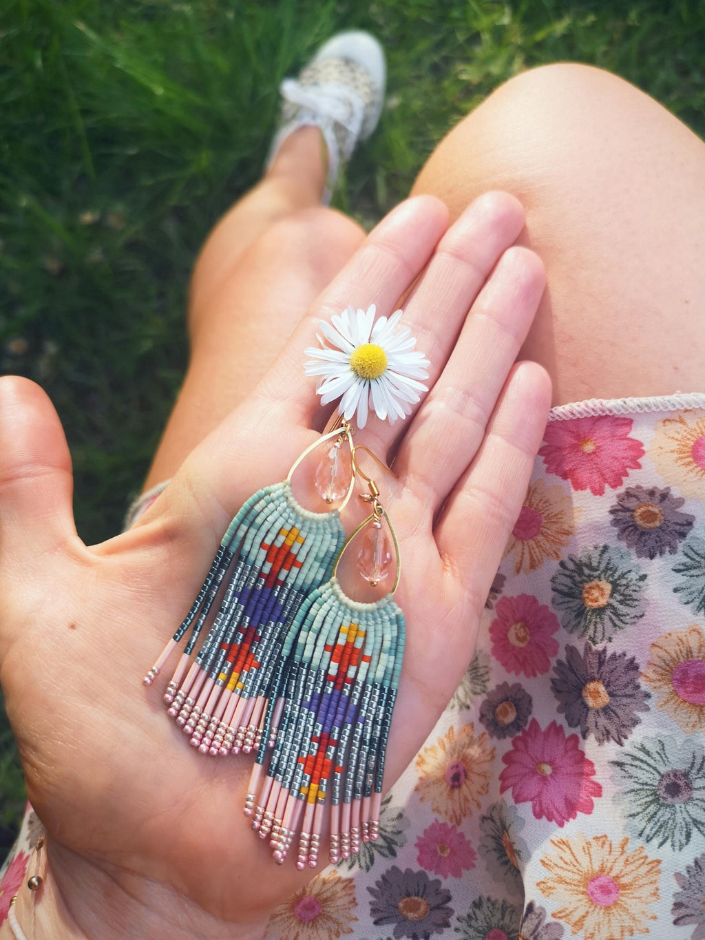 Peut inclure: Boucles d'oreilles en perles faites à la main, en forme de goutte d'eau et à franges. Les boucles d'oreilles présentent une palette de couleurs bleu clair et rose avec des accents orange, rouge et violet. Une main tient les boucles d'oreilles avec une marguerite.